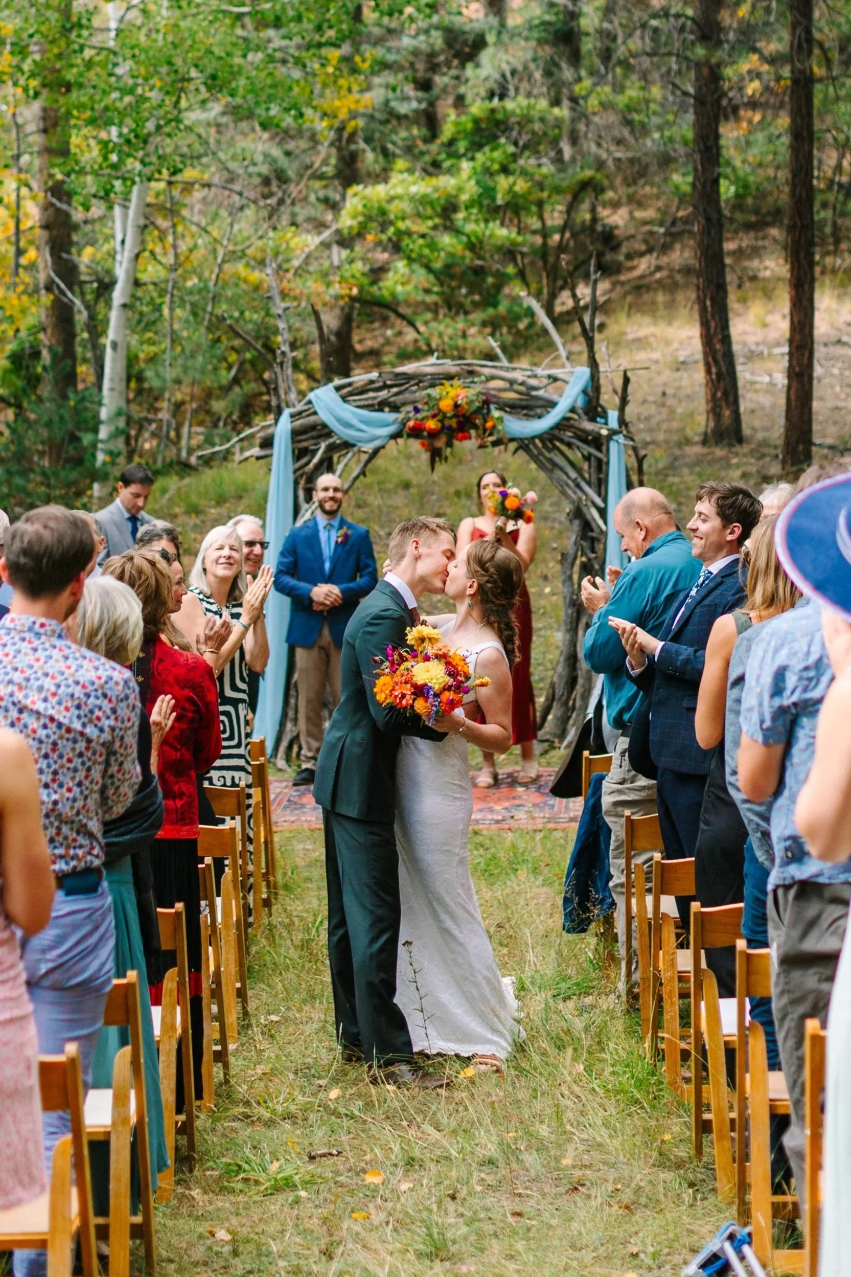 A wedding ceremony outdoors in a wooded area with a bride and groom kissing, surrounded by guests. The altar is decorated with flowers and light blue fabric, and the guests are standing and clapping.