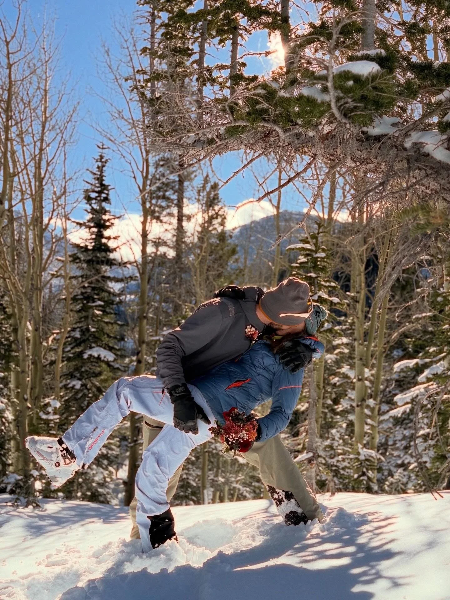 Two people, dressed in winter clothing, are snowshoeing in a snowy forest surrounded by tall trees, with a mountain in the background, during sunset or sunrise.