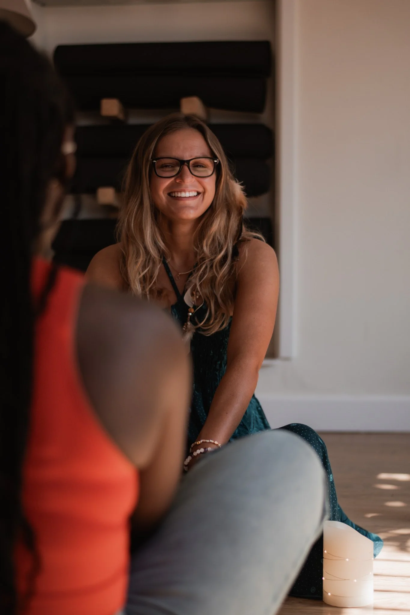 A woman with long wavy hair, glasses, and a black necklace smiling and looking at someone. She is indoors with a dark shelf or furniture behind her and a white wall. Part of another person wearing an orange top is visible in the foreground.