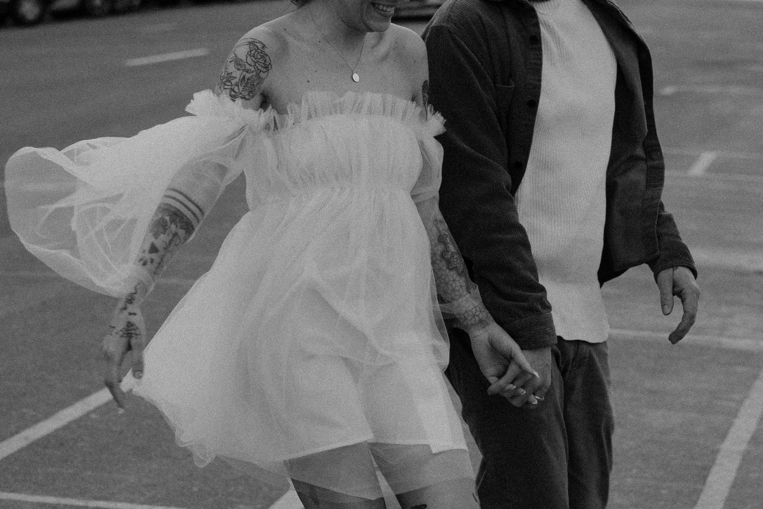 A couple holding hands while ice skating, the woman is wearing a white dress with off-shoulder ruffle sleeves and tattoos on her arms, the man is wearing a dark jacket and light-colored shirt, both smiling.