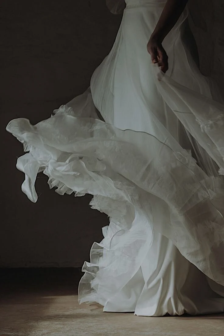 Close-up of a woman in a flowing white wedding gown, standing on a wooden floor with a dark background.