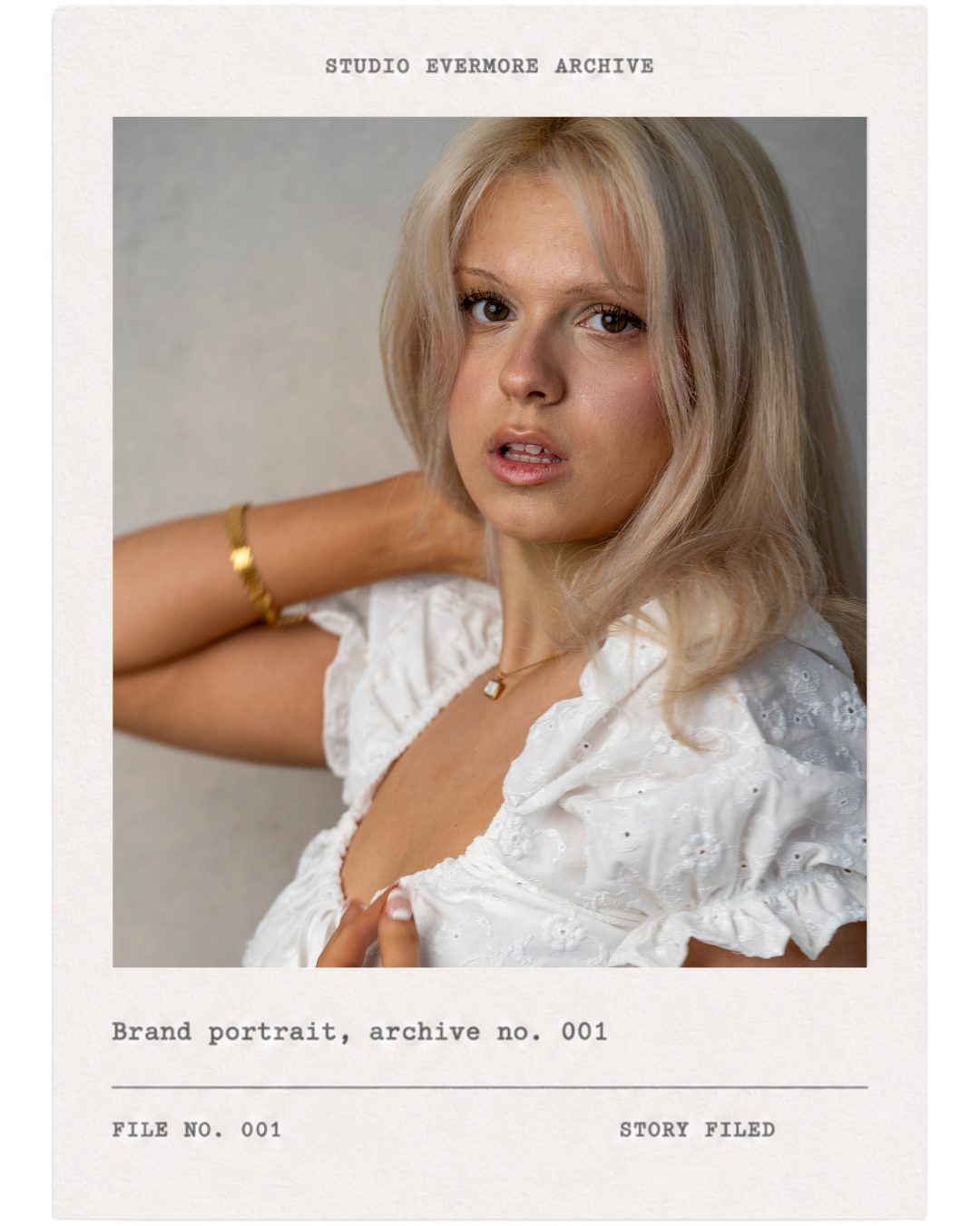 A woman with shoulder-length blonde hair and light skin posing in a white eyelet blouse, wearing a gold bracelet and necklace, with a neutral background.