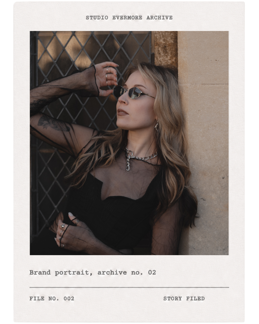 A woman with blonde hair and sunglasses posing against a stone wall and metal gate, wearing black clothing and silver jewelry.