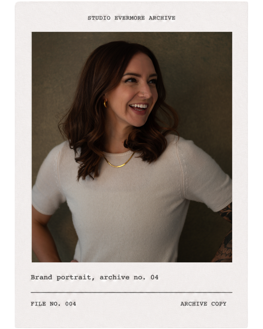 A smiling woman with brown hair, gold jewelry, wearing a white top, standing against a neutral background.