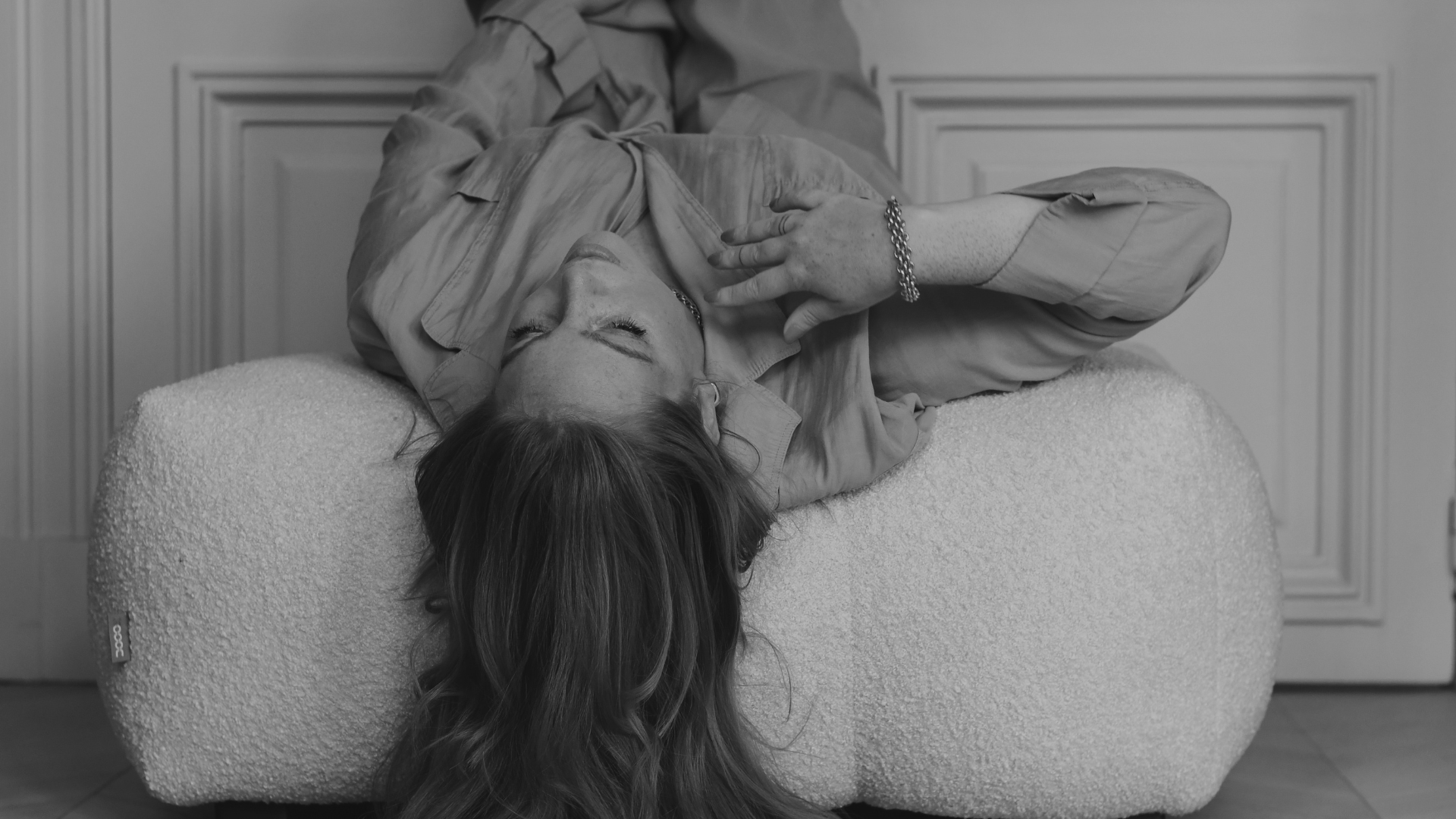 A woman with long hair lying on a textured ottoman with her eyes closed and hand on her chest, inside a room with paneled walls.