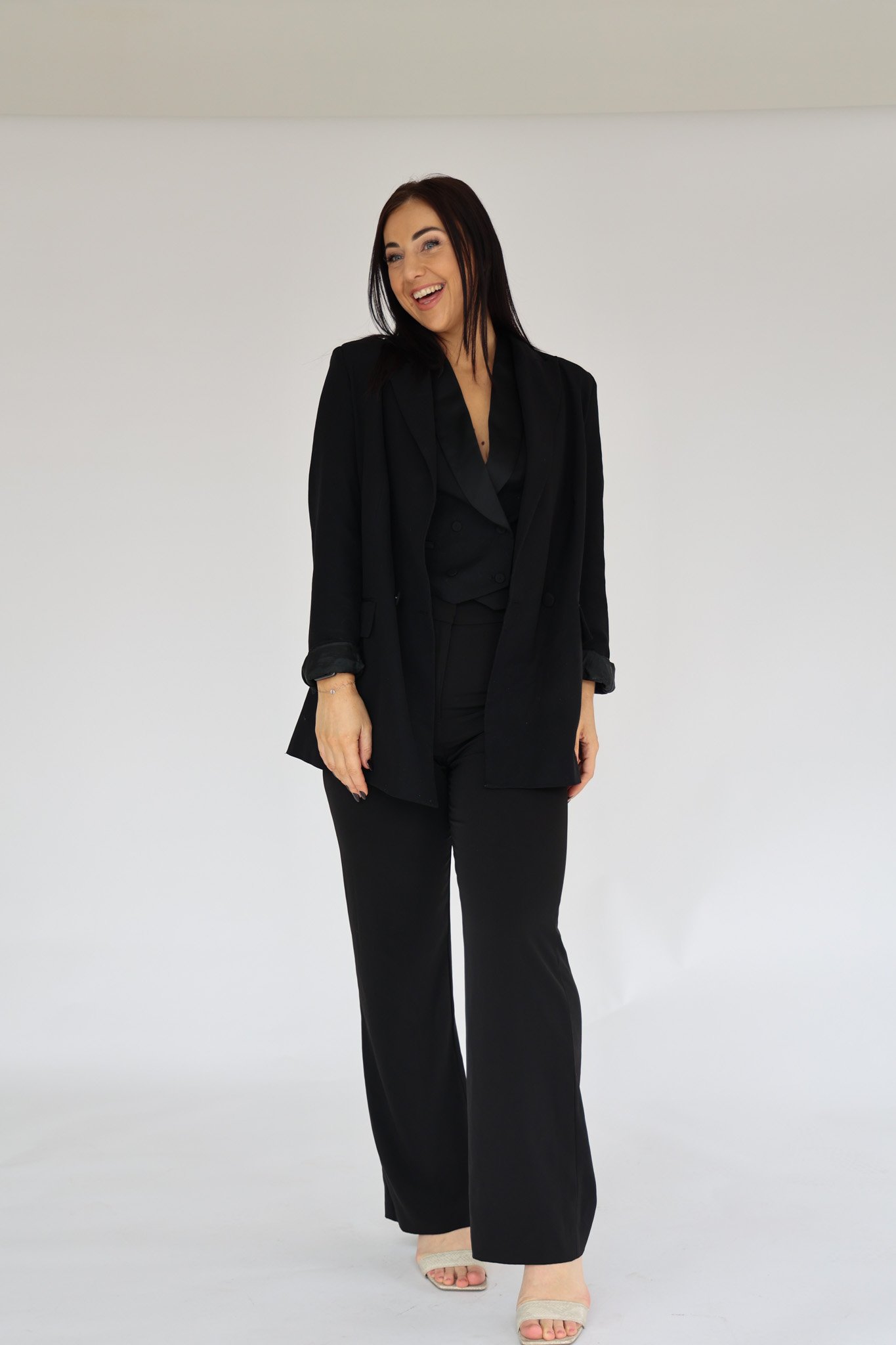 A woman with long dark hair wearing a black blazer, black pants, and beige high heels, smiling and looking to the side, standing against a plain white background.