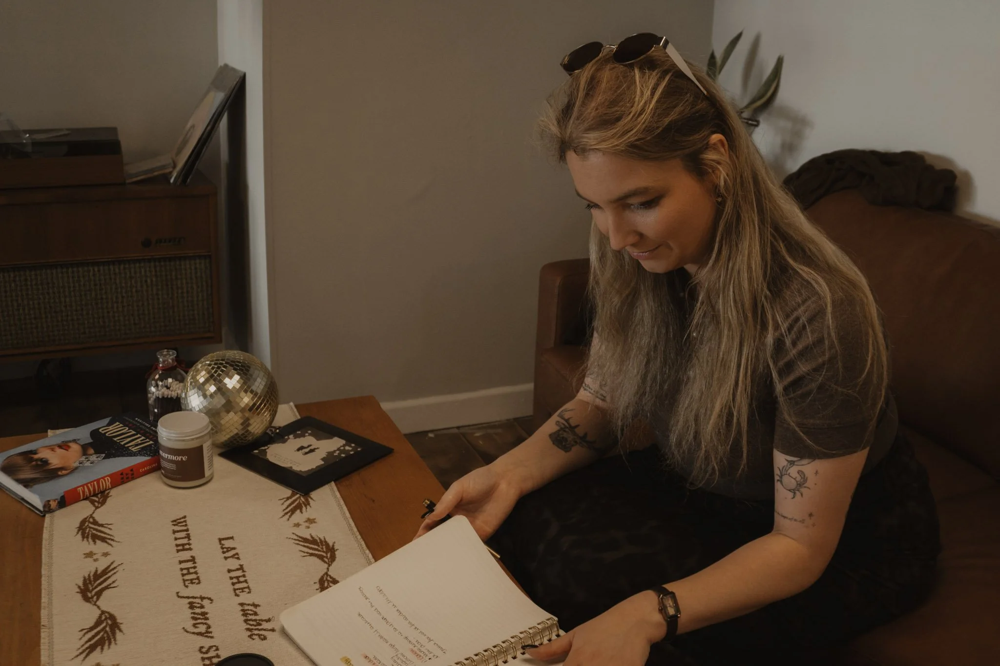 A woman with long blonde hair, tattoos on her arms, and sunglasses on her head, is sitting at a table with various objects including books, a notebook, and a decorative gold sphere. She is looking down at her hand and smiling.