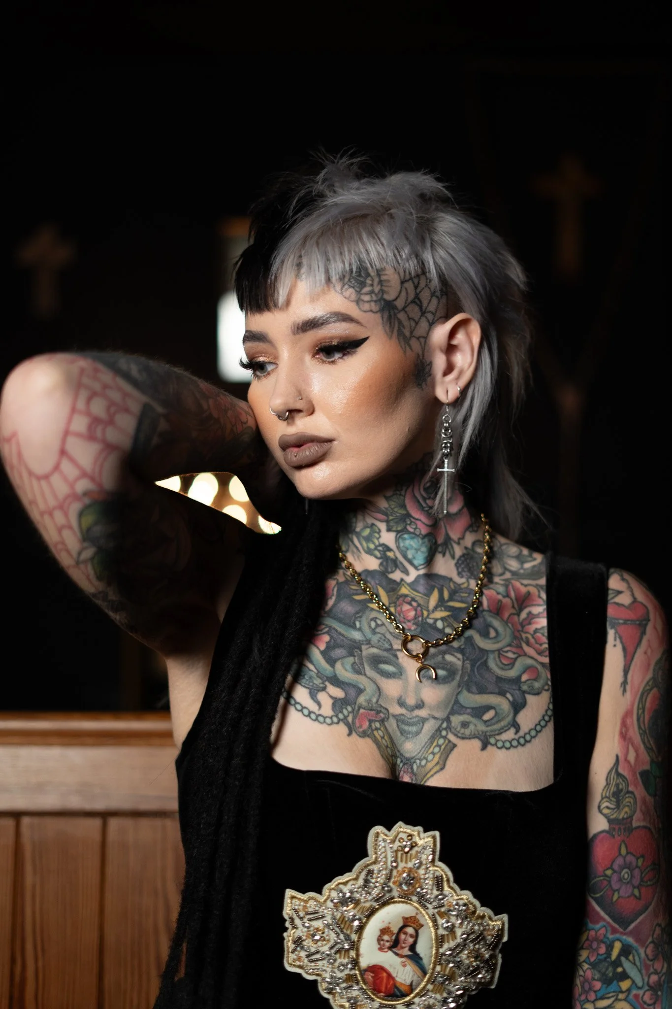 A woman with tattoos, piercings, and short, black and gray hair, wearing earrings, necklaces, and a black outfit, standing indoors with wooden paneling and religious crosses visible in the background.
