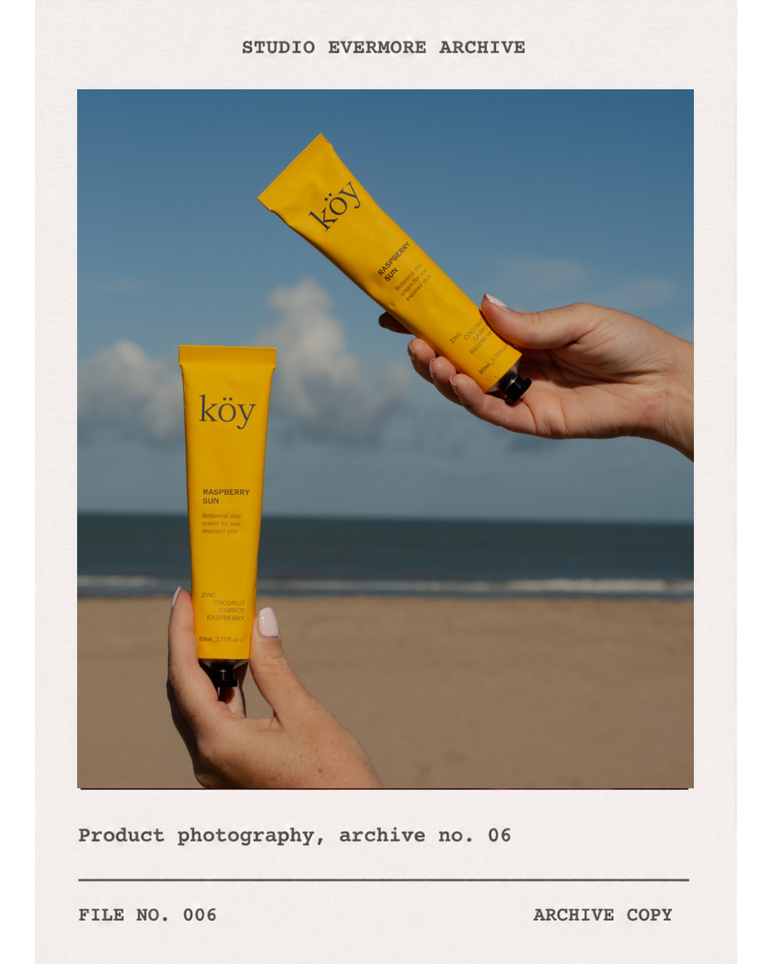 Two hands holding yellow tubes of Köy Raspberry Sun sunscreen lotion against a beach backdrop with blue sky and clouds.