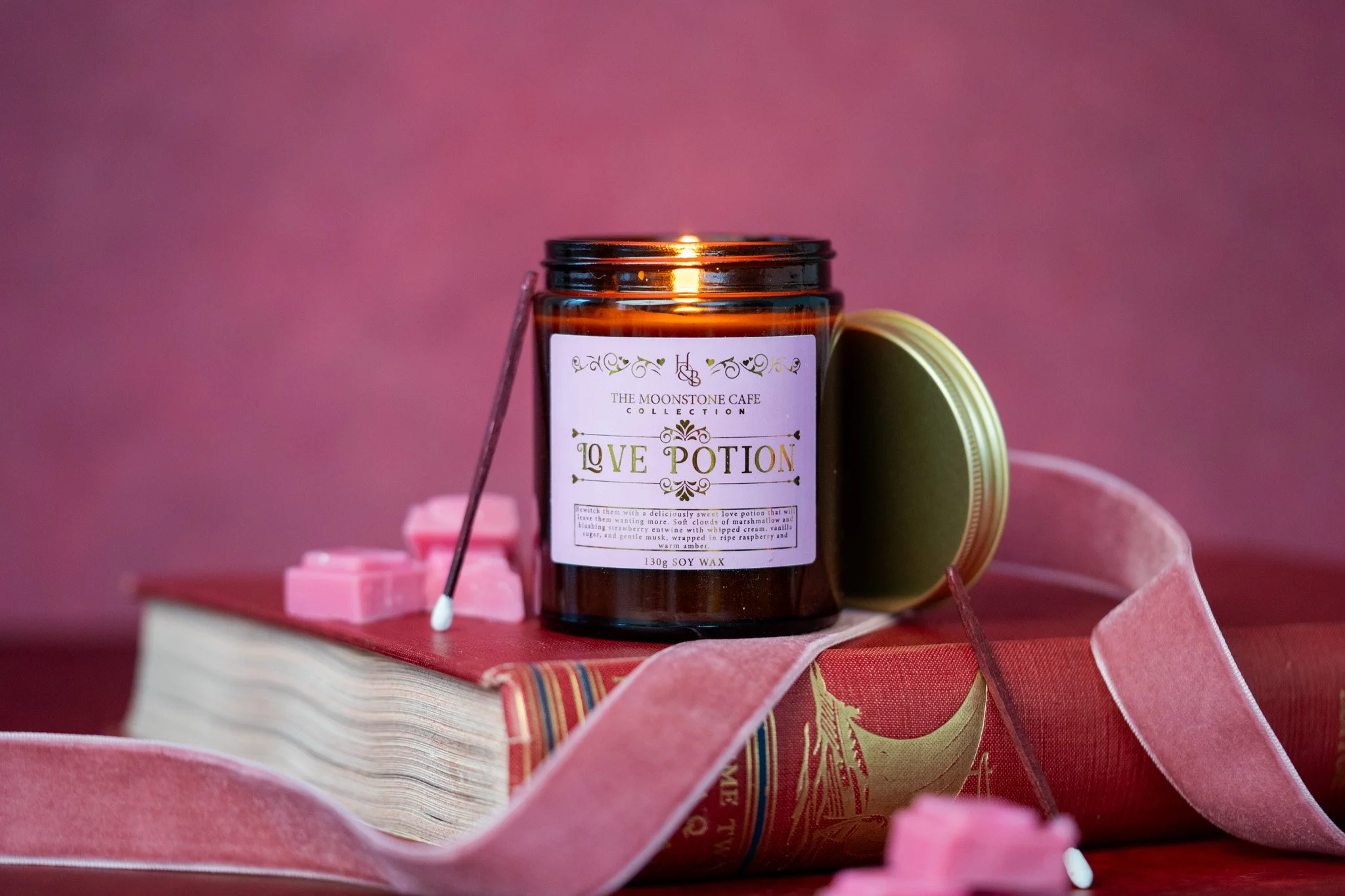 A brown jar labeled 'Love Potion' from The Moonstone Cafe Collection, surrounded by pink and red decorative items, placed on a pink surface with a pink background.