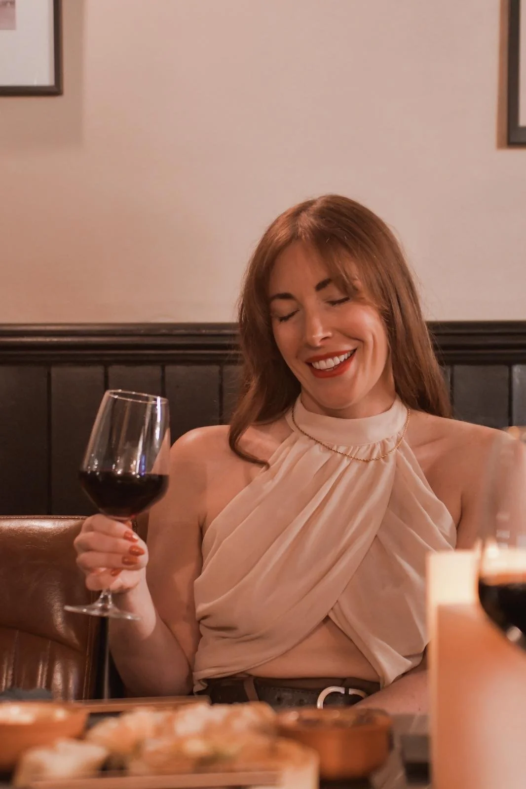 A woman with red hair and a beige halter top smiling and holding a glass of red wine at a restaurant.