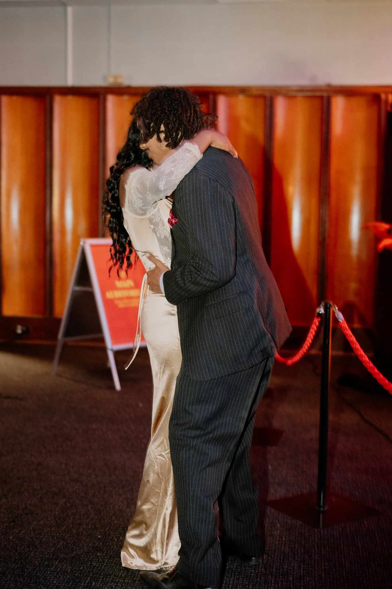 A man in a dark pinstripe suit and a woman in a light satin dress are hugging in a warmly lit indoor setting with wooden paneling in the background.