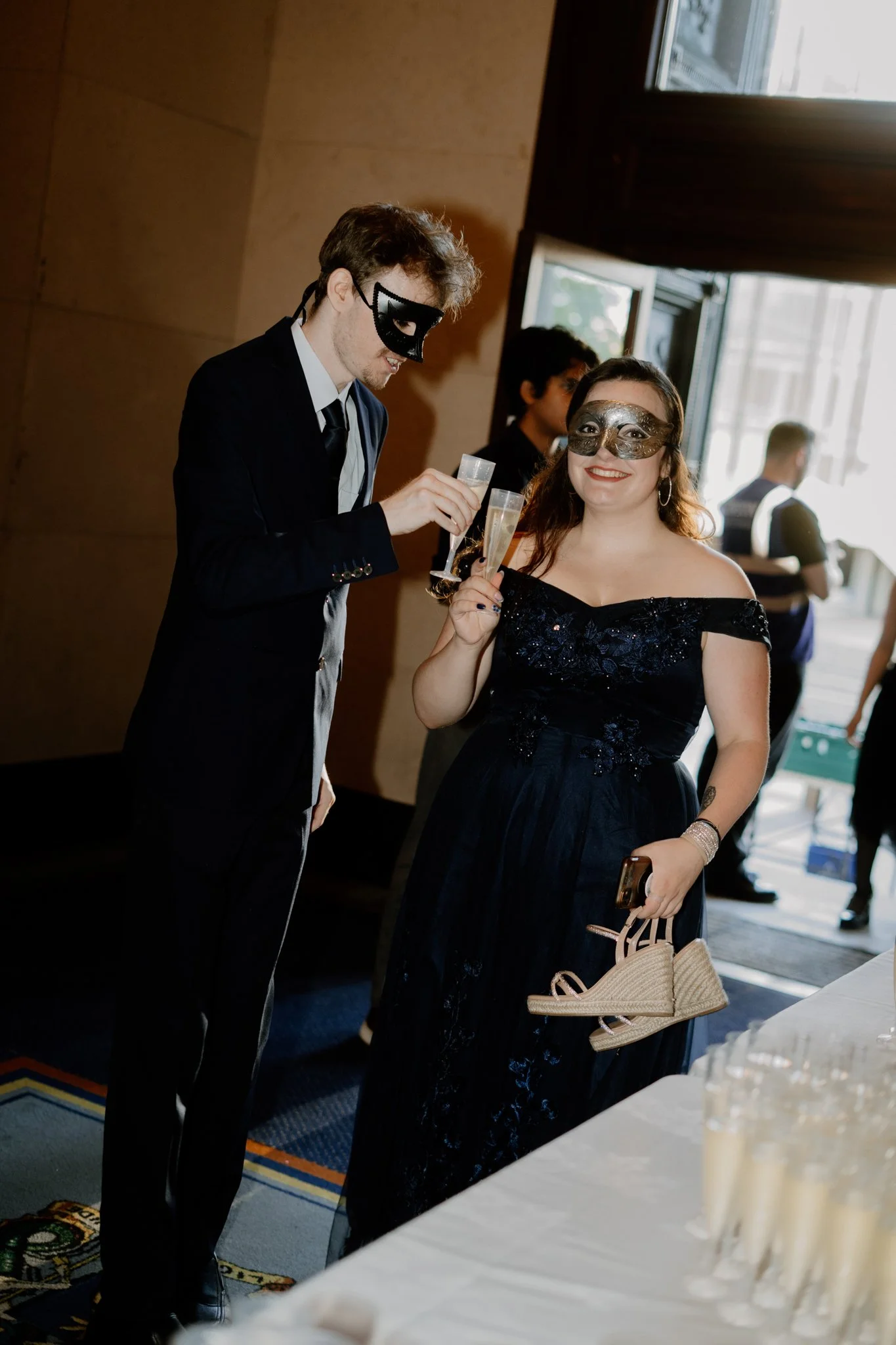 A man and a woman dressed formally, both wearing masks, holding glasses of champagne at a celebration or party. The woman is smiling and holding a beige handbag.