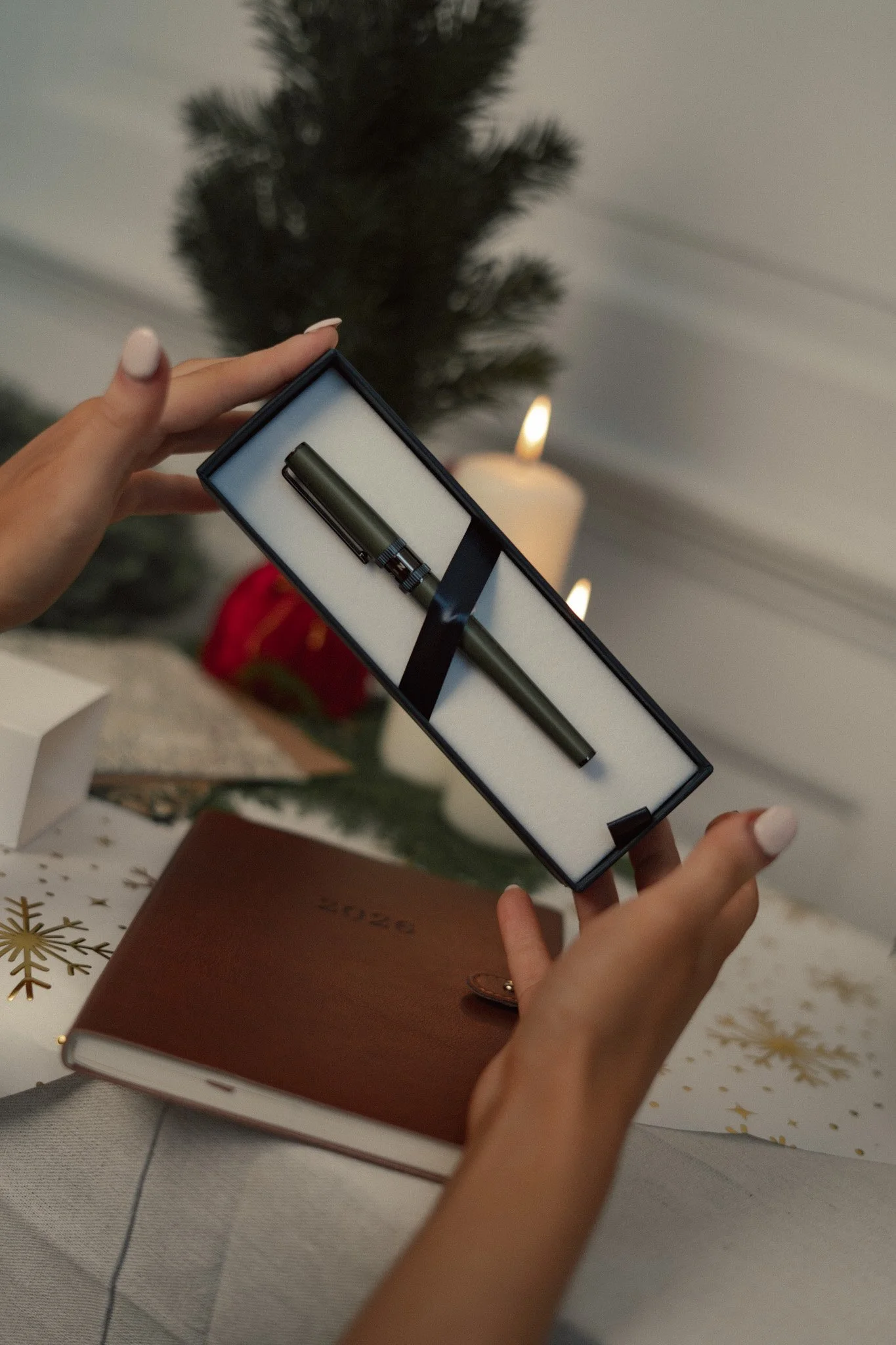 Person holding a gift box with a pen inside, with a Christmas candle and a decorated tree in the background.