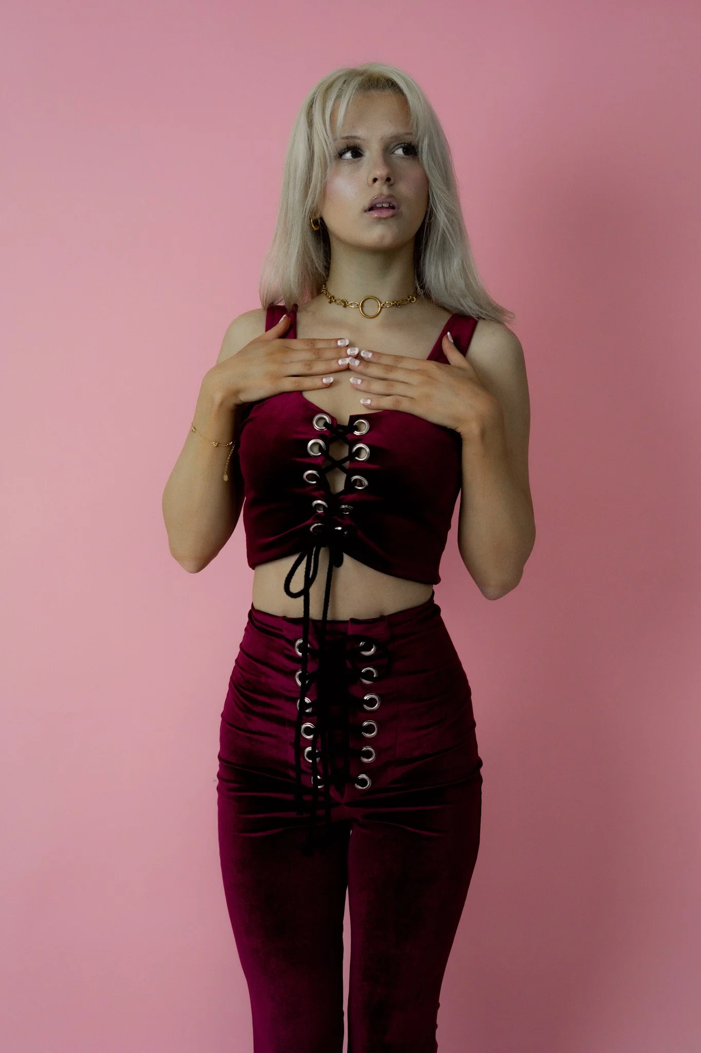 A woman with platinum blonde hair in a burgundy organdy crop top and matching skirt with black lace-up details, standing against a pink background.