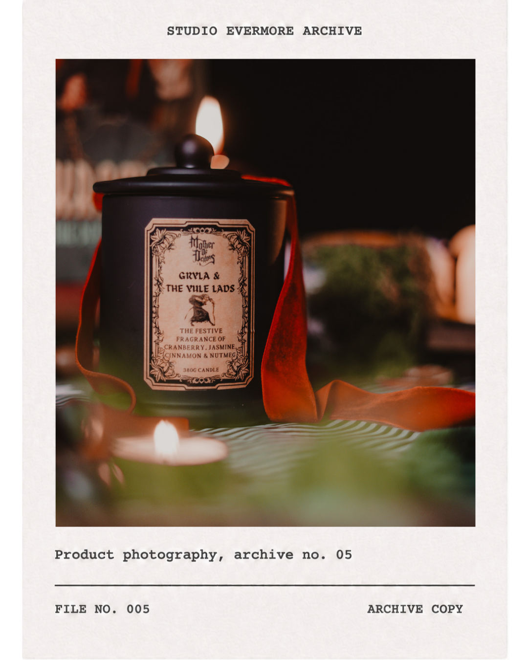 Close-up of a black candle jar with a vintage-style label, surrounded by a red ribbon and a small candle flame in the foreground, on a green tablecloth, with a blurred background.