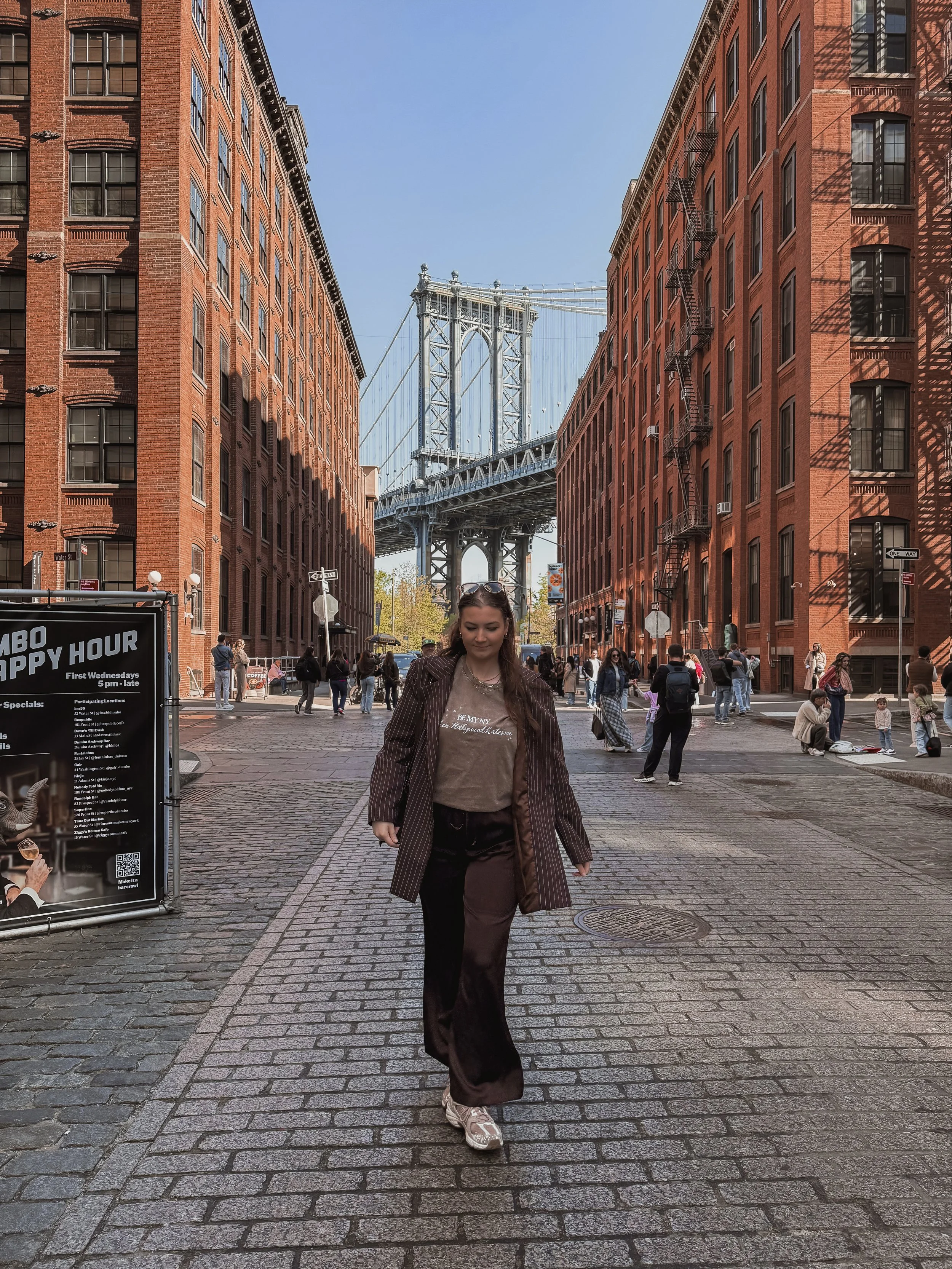 A young woman walking on a cobblestone street in a city, with the Brooklyn Bridge visible in the background, flanked by brick buildings on either side.
