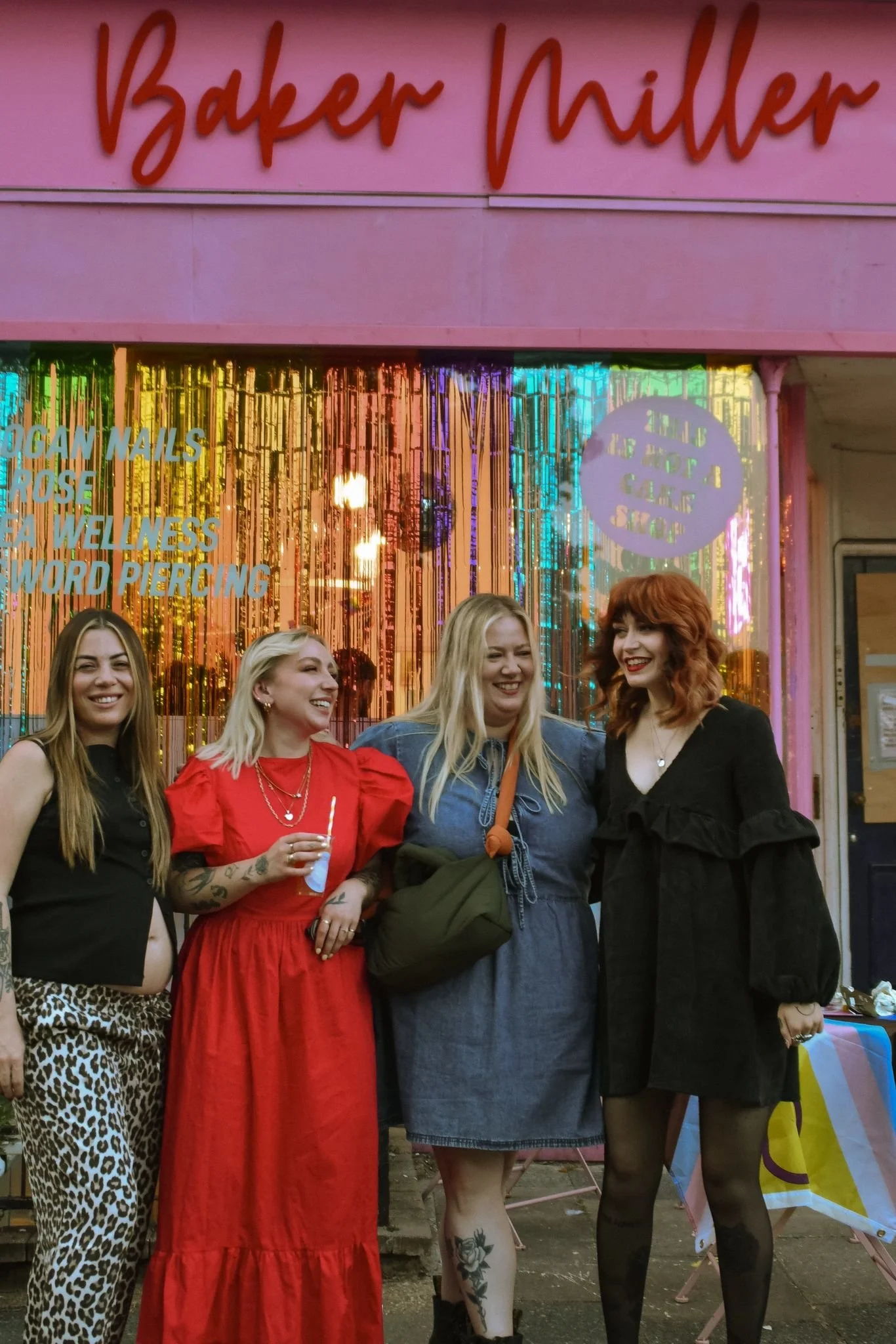 Four women standing together in front of a pink storefront with colorful metallic curtains. They are smiling and appear happy, dressed in various outfits including animal print pants, a red dress, a denim dress, and a black dress. One woman is holding a drink.