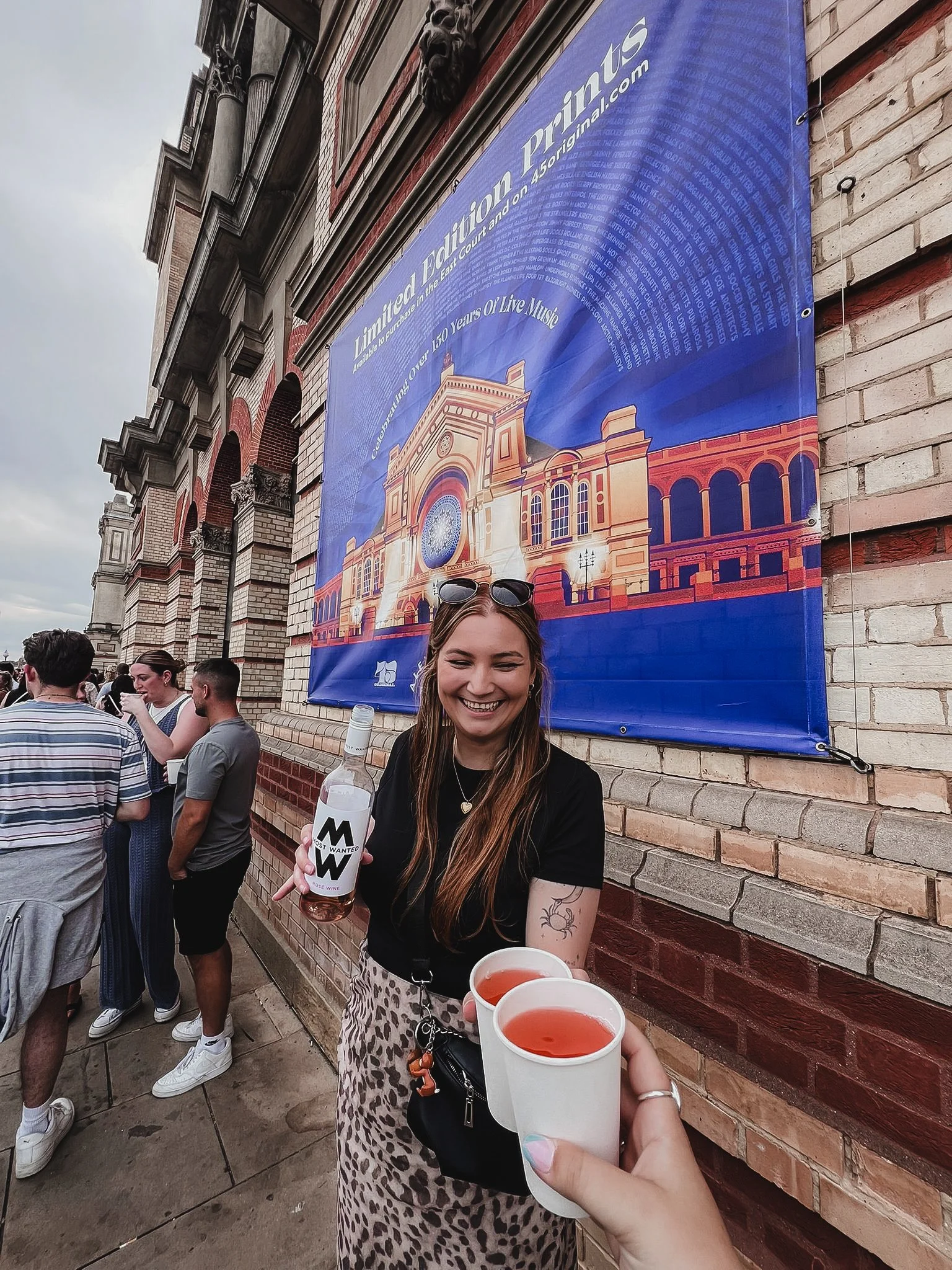 A woman smiling and holding three cups of a pink beverage at an outdoor gathering next to a brick wall and a large blue banner. The girl in the foreground is wearing a black shirt, leopard print pants, and sunglasses on her head. There are other people in the background standing and talking.