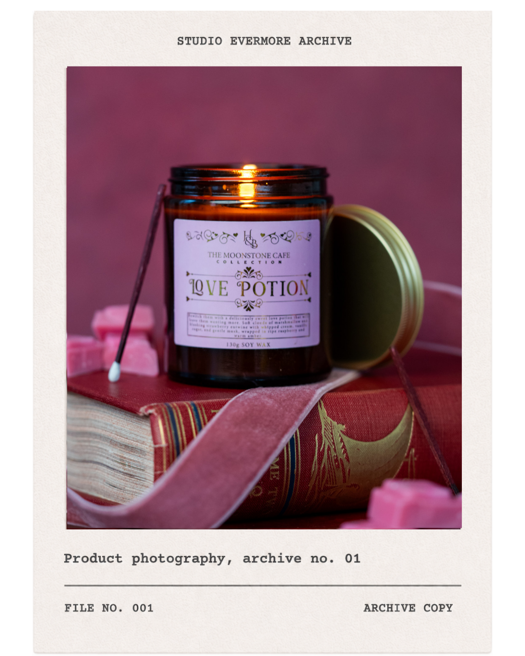 A brown jar labeled "Love Potion" from The Moonstone Cafe Collection, with a gold lid, resting on a vintage book with pink sweets, a cotton swab, and pink flowers around it.