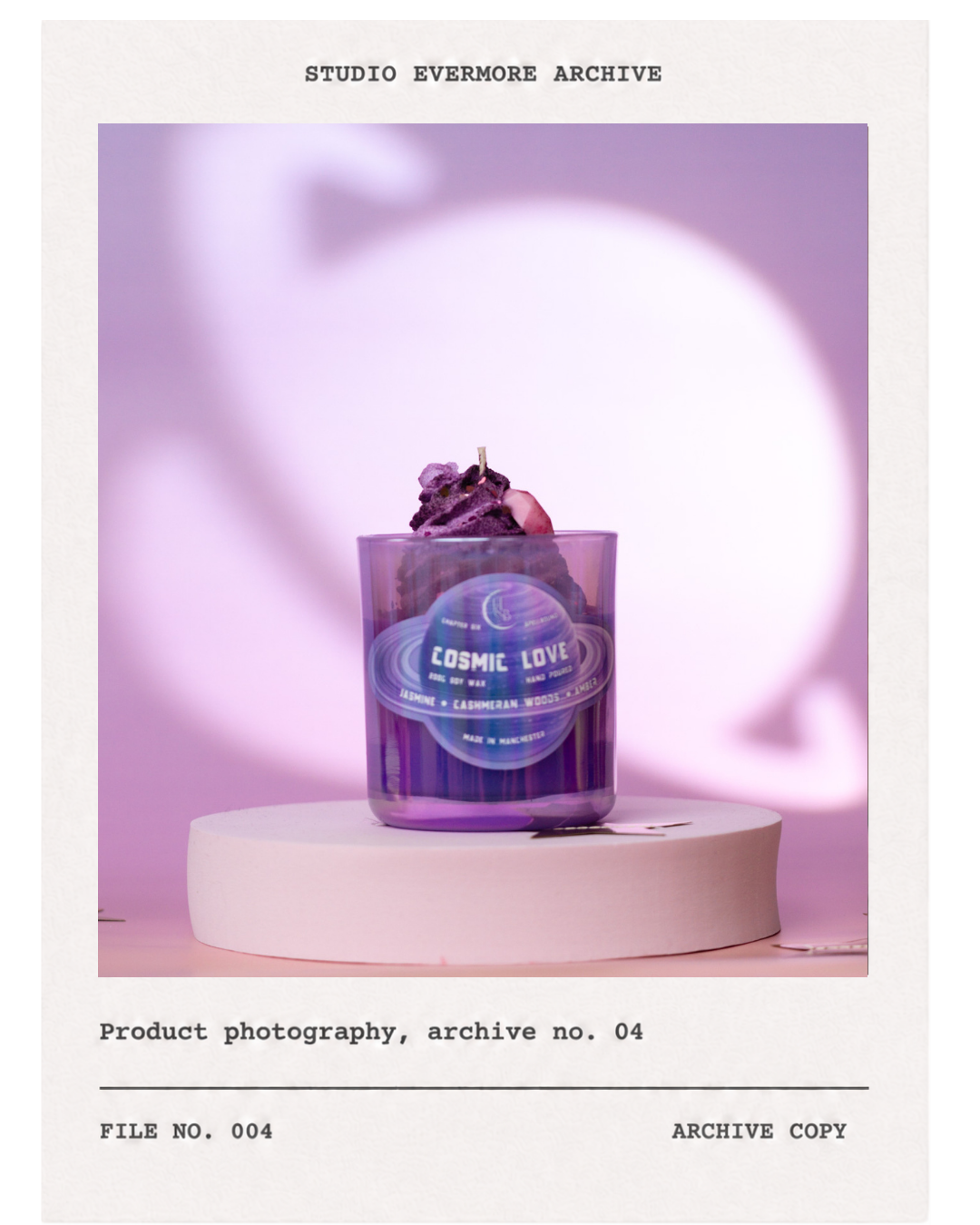 Photograph of a purple candle with a label 'Cosmic Love' on a white pedestal, with makeup on top, against a purple background.