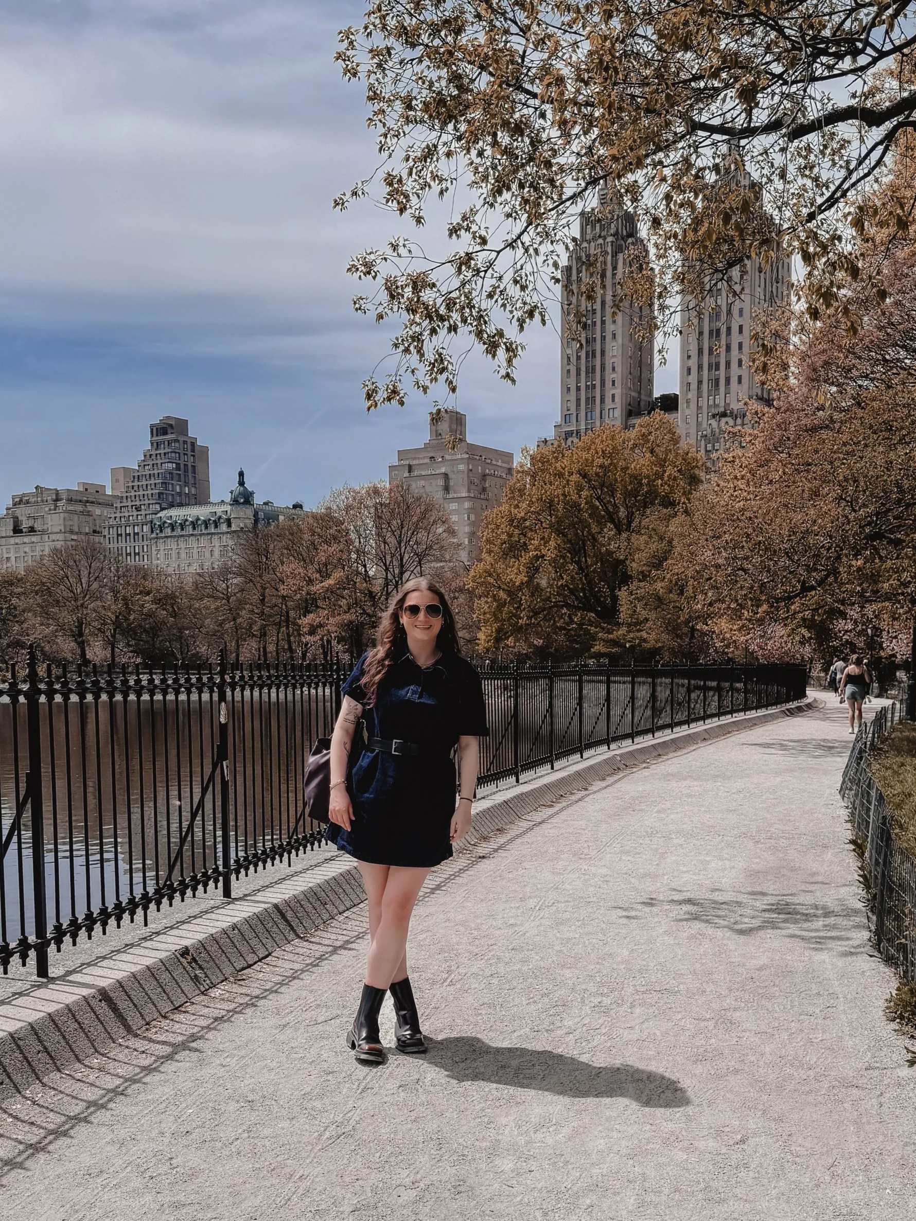 A young woman in a black dress and black boots standing on a paved pathway near a river, with trees in full bloom and tall buildings in the background under a partly cloudy sky.