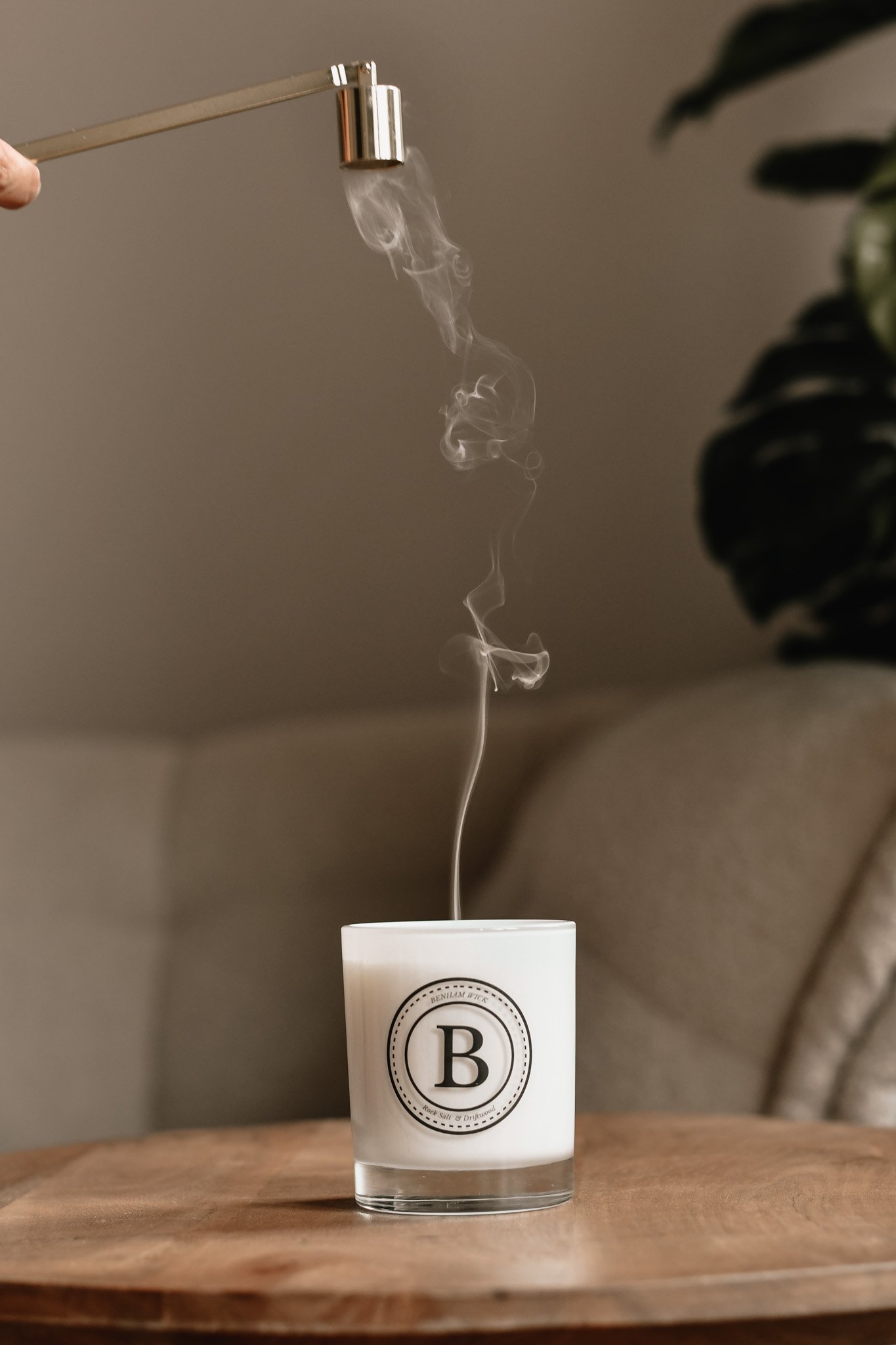 A white candle with a black logo and letter B on it, with smoke rising from its wick, on a wooden surface. A blurred plant is on the right side of the image.