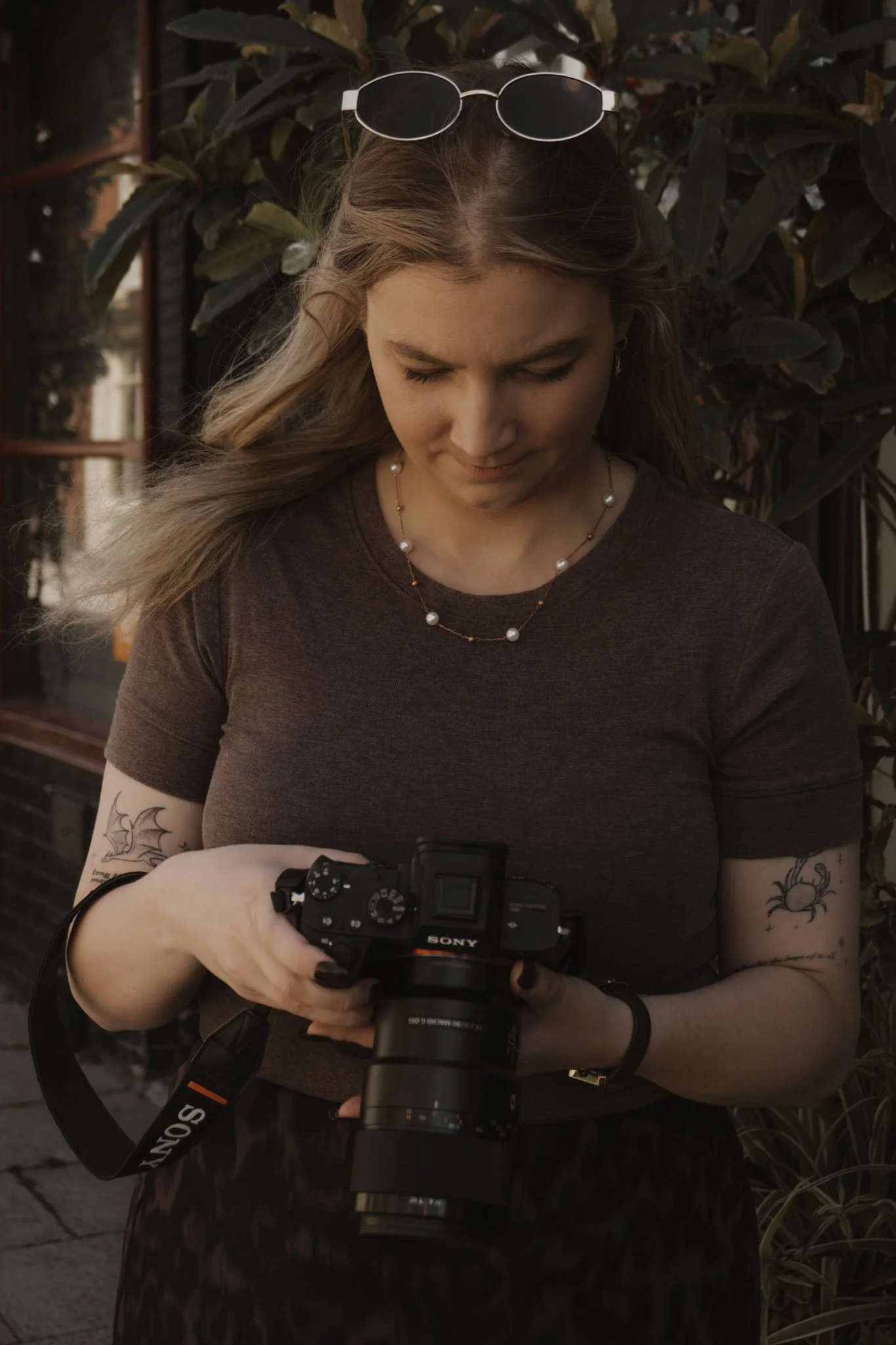 A woman with long hair, sunglasses on her head, and tattoos on her arms, wearing a dark t-shirt and pearl necklace, is looking down at a Sony camera she is holding in her hands. She is outdoors in front of leafy plants.