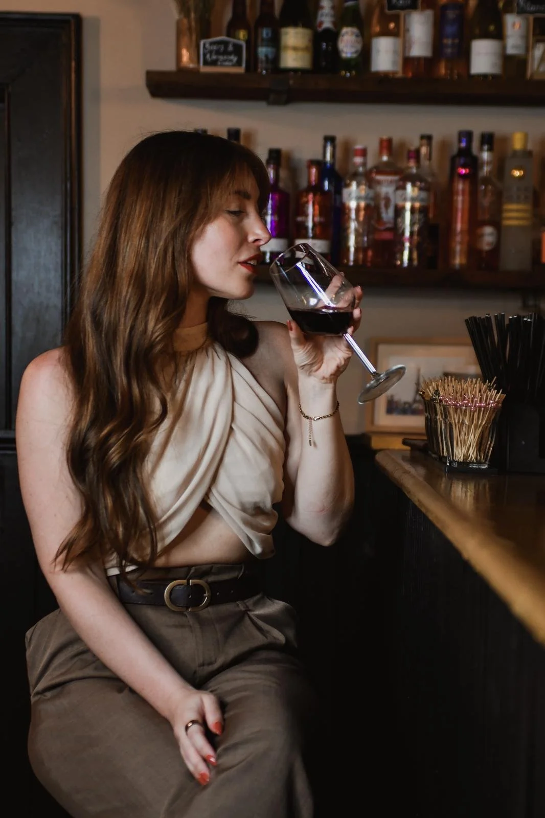 A woman with long brown hair drinking red wine in a bar, standing near a counter with bottles of liquor in the background.