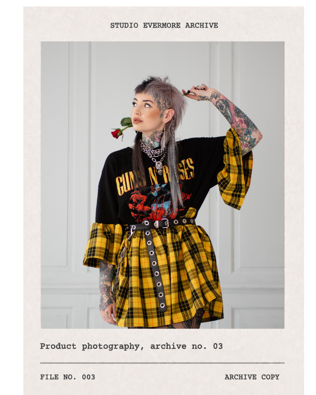 A woman with tattoos and short, spiky hair holding a red rose in her mouth, wearing a black Guns N' Roses t-shirt layered with a yellow plaid skirt and matching sleeve, accessorized with chains and a belt, standing against a white wall.