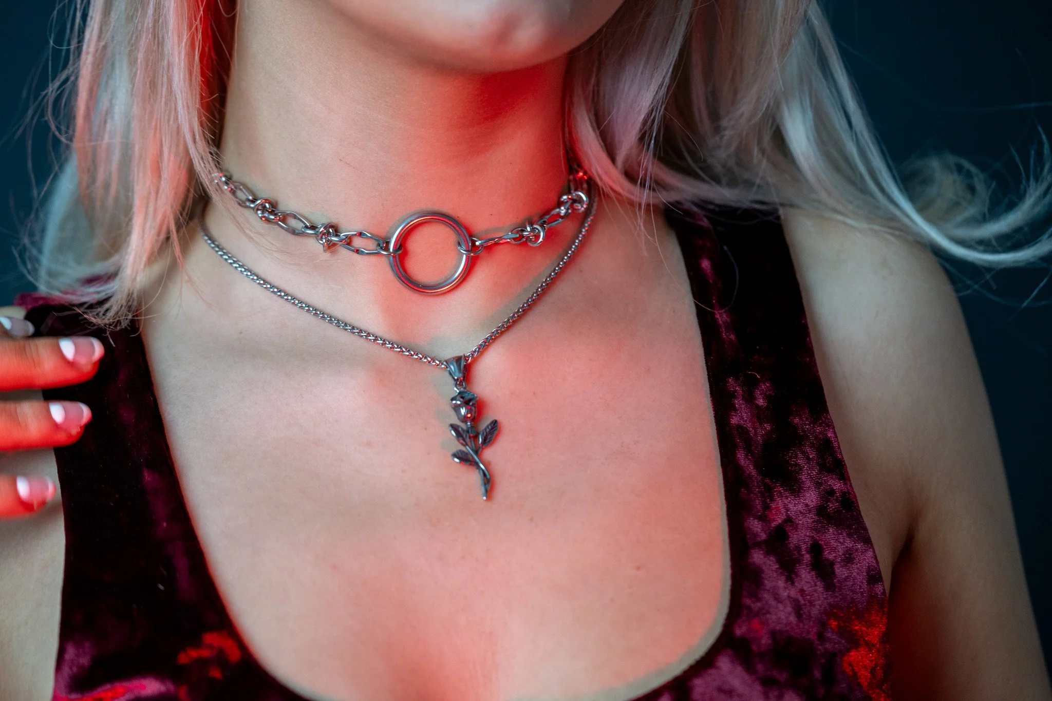 Close-up of a woman's neck and chest showing layered silver necklaces, including a chain with a large circular ring, a thin chain with a pendant, and a decorative necklace with a leaf design. She is wearing a dark, velvety, floral-patterned top or dress, and has long blonde hair.