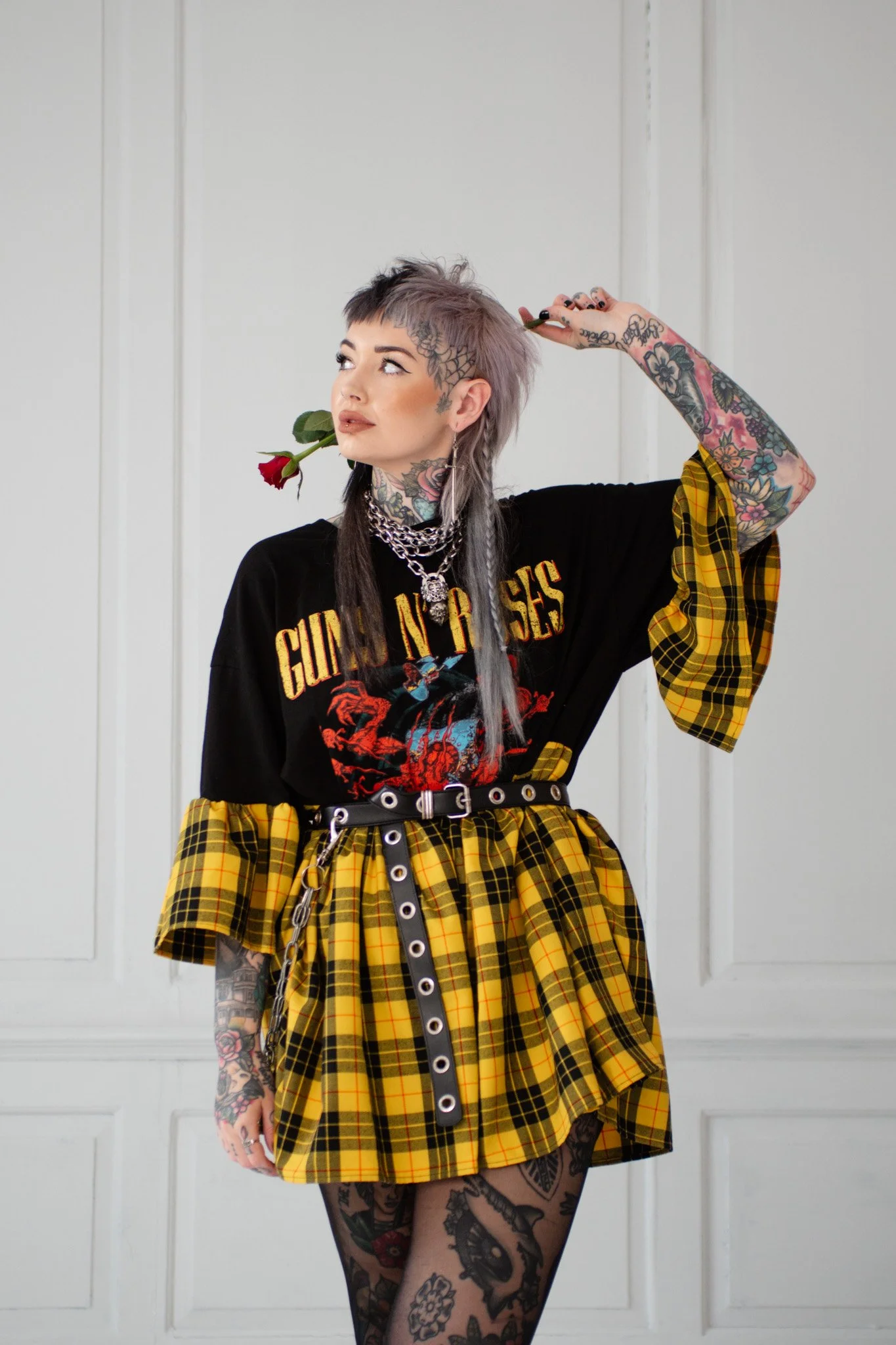 A woman with short, purple-dyed hair and tattoos, holding a red rose in her mouth, wearing a Guns N' Roses t-shirt, plaid yellow skirt, and multiple jewelry pieces, standing in front of a white paneled wall.