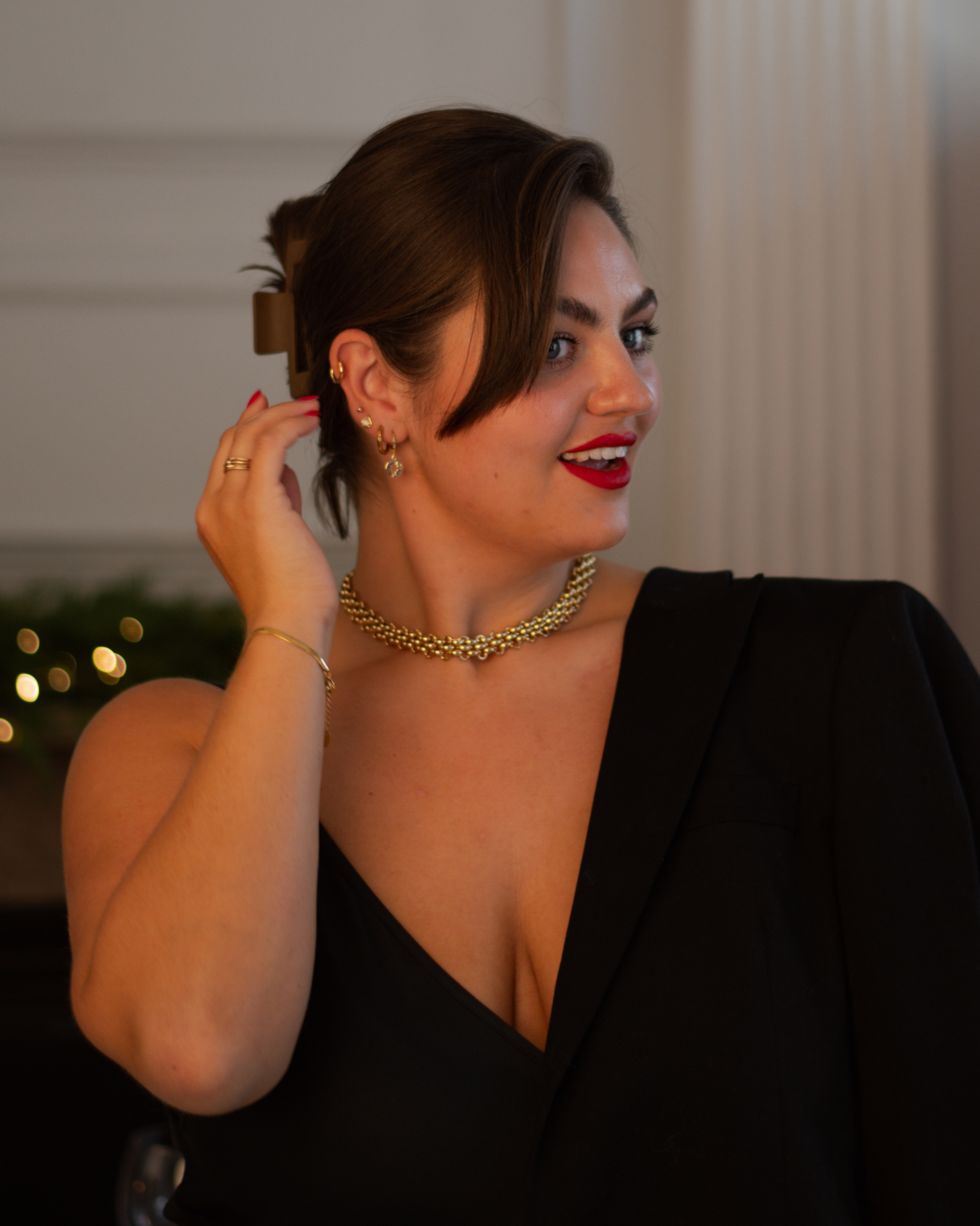 A woman with short brown hair wearing a black dress, gold jewelry, and red lipstick, adjusting her hair in a formal indoor setting.