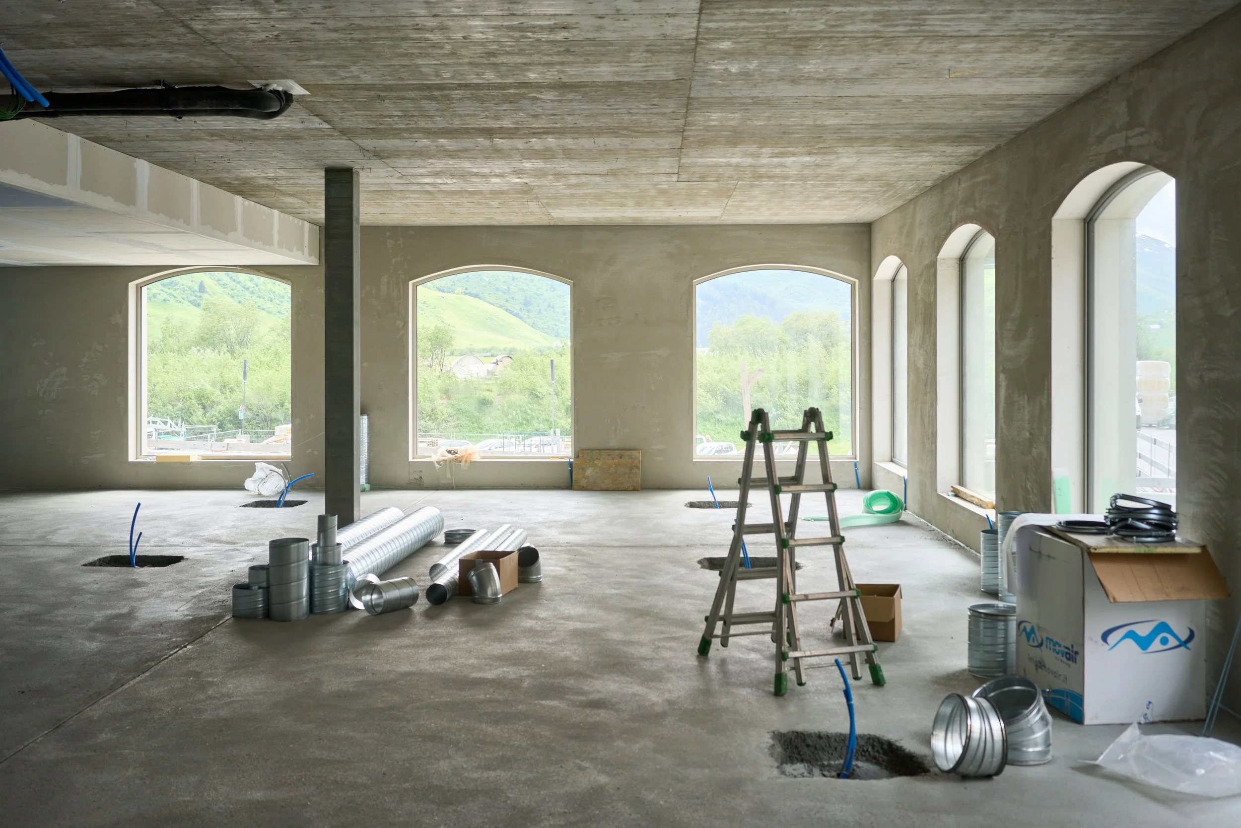 Interior view of a building under construction with large arched windows, construction materials, a ladder, and construction pipes on the concrete floor, with a scenic landscape visible through the windows.