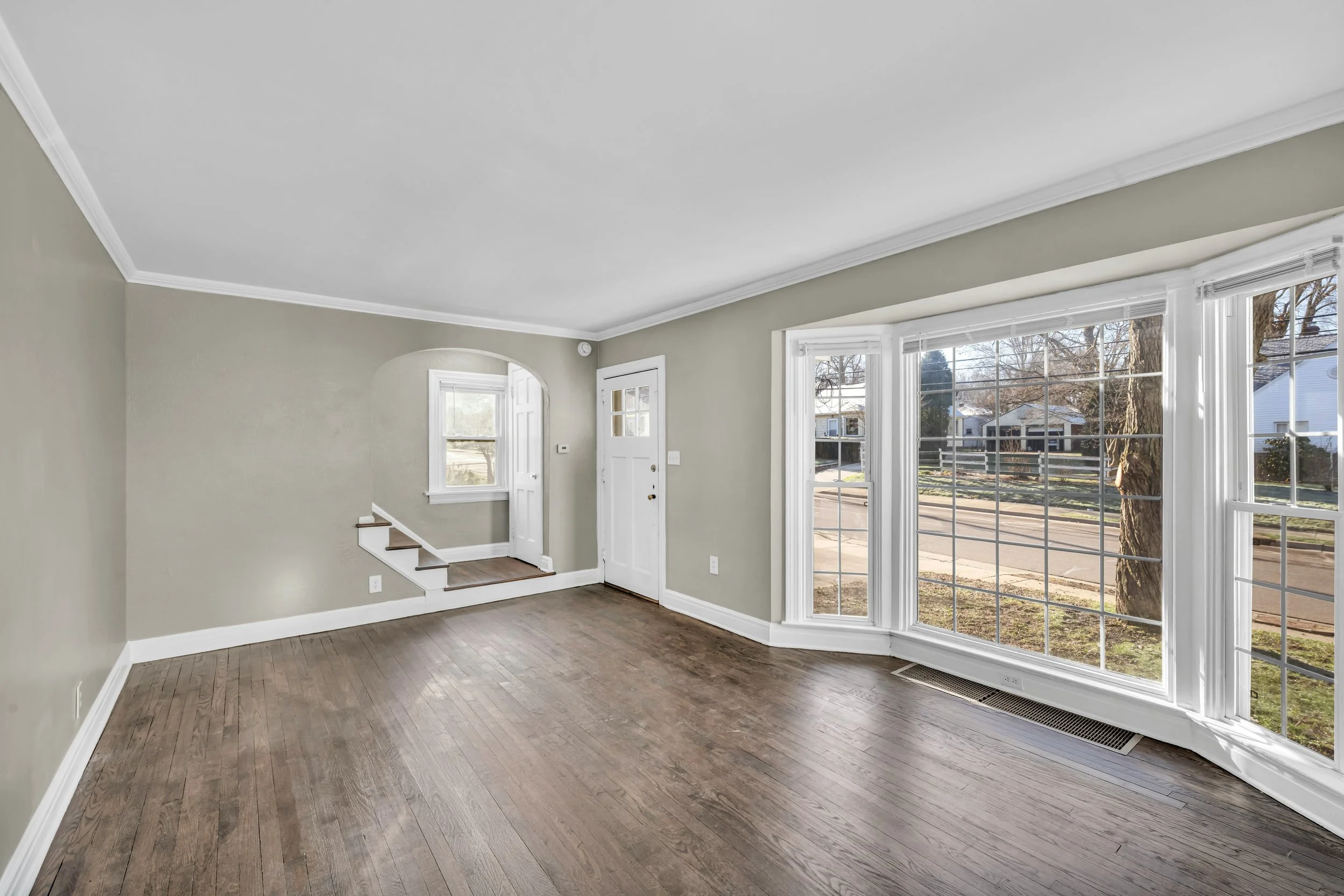 Bright living room with large bay windows, hardwood floors, pale green walls, white trim, and a staircase near the entrance.