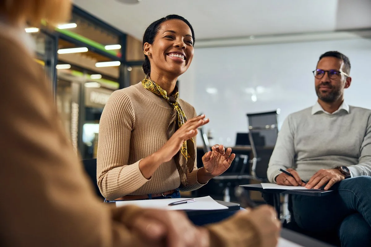 A woman smiling and talking during a meeting with two other people in an office setting.
