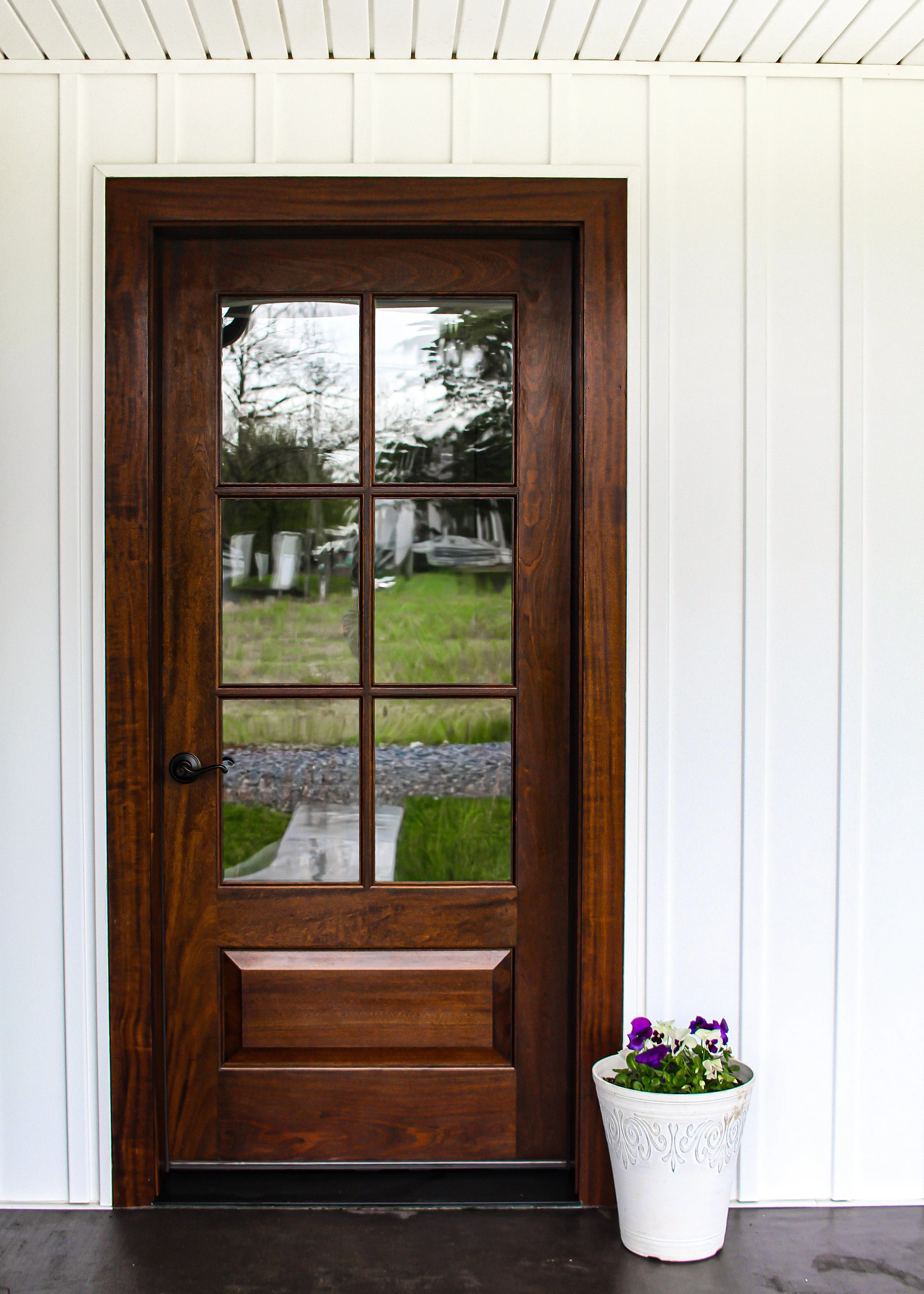 A wooden door with glass panels, installed in a white wall made of vertical siding. Outside, a garden with trees and grass, with a walkway visible through the glass. A white decorative planter with purple and white flowers is positioned to the right of the door inside.