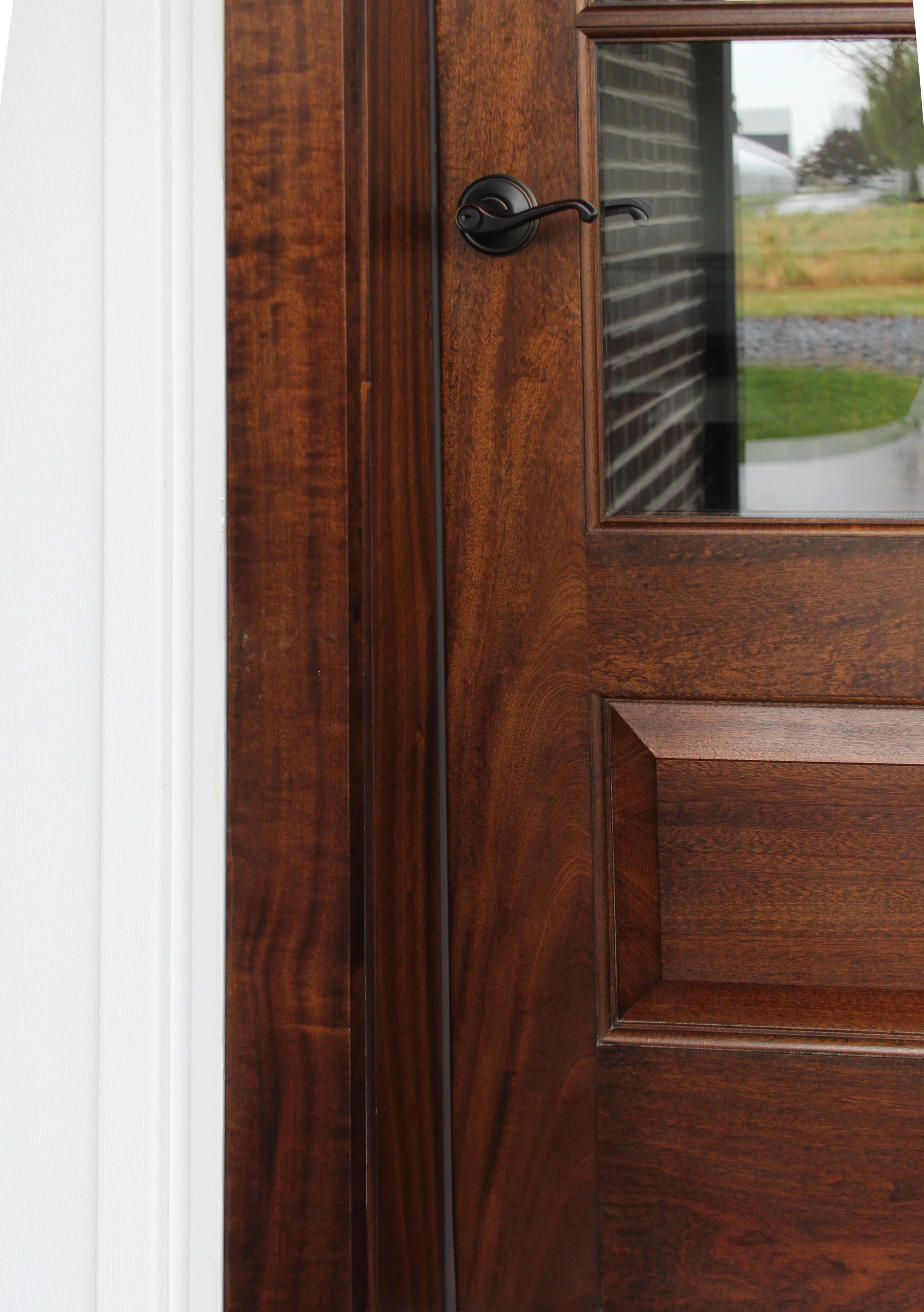 A close-up of a wooden door with a black handle and a glass window, showing a brick wall and some greenery outside.