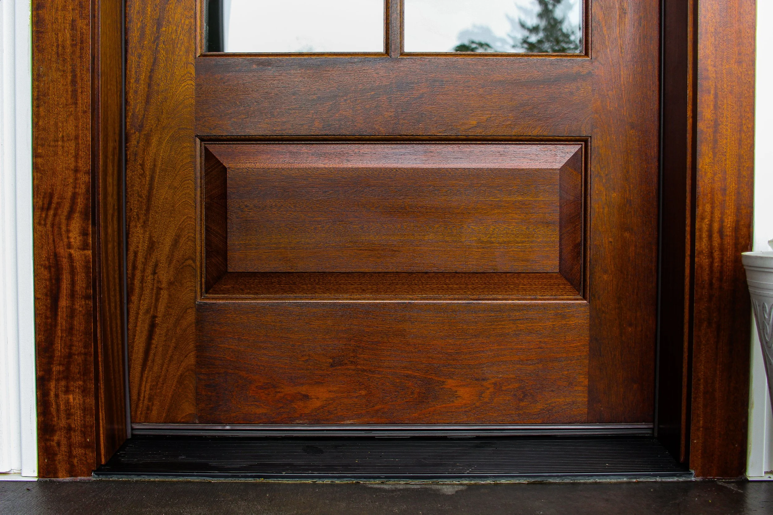 Close-up of a wooden front door with glass window panes at the top, surrounded by matching wood trim and a black rubber mat at the threshold.
