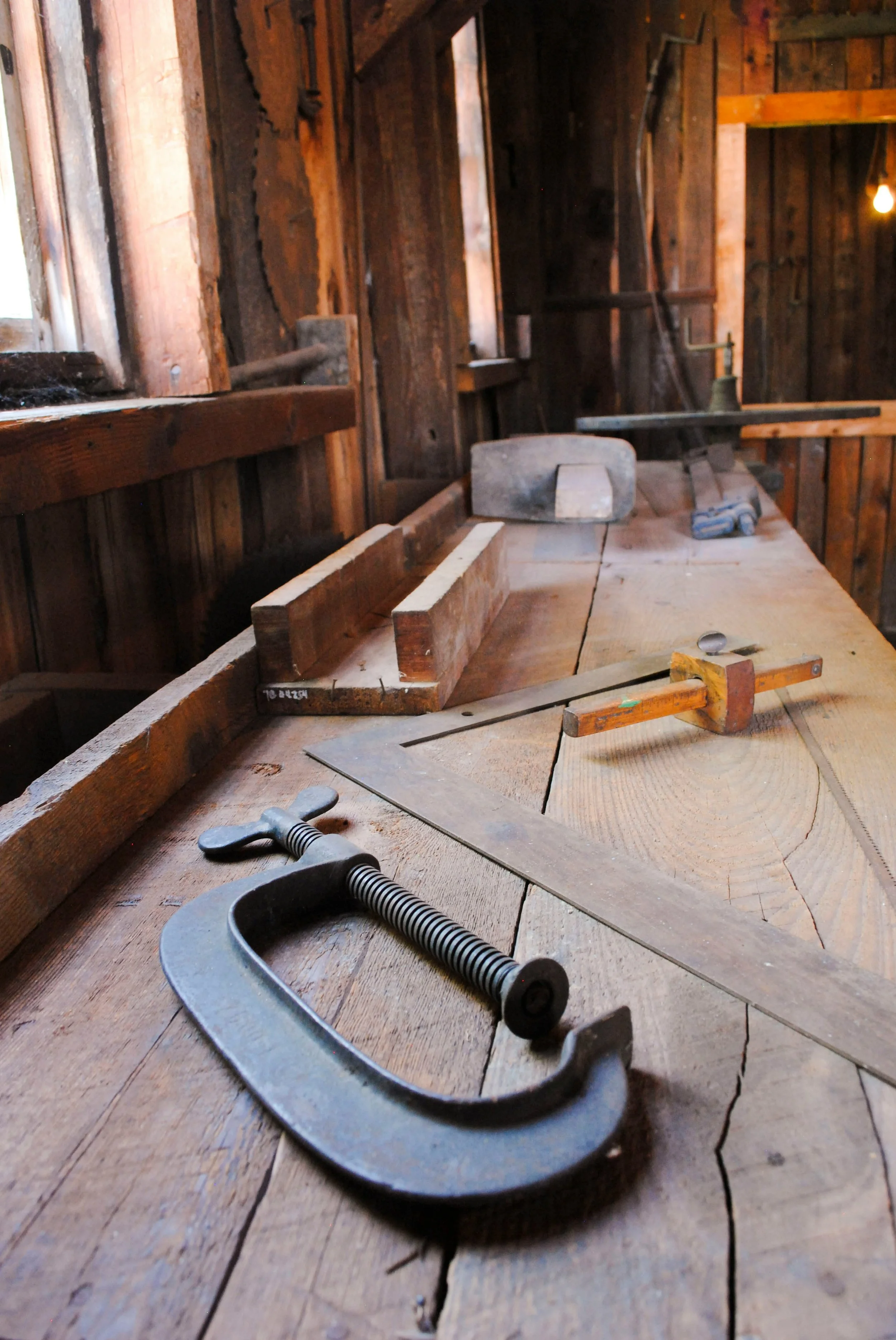 Inside a rustic woodworking shop, a workbench with woodworking tools including a C-clamp and handsaw, with a wooden bench and various hand tools in the background.