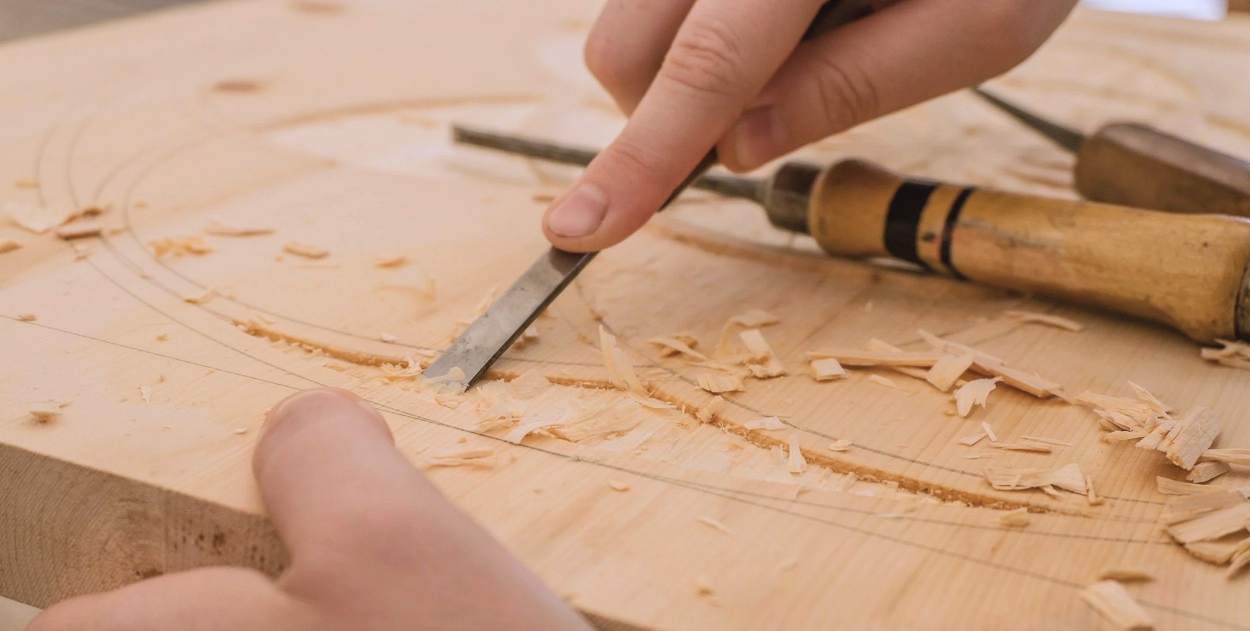 Person carving wood with a chisel and mallet on a wooden workbench, wood shavings scattered around.