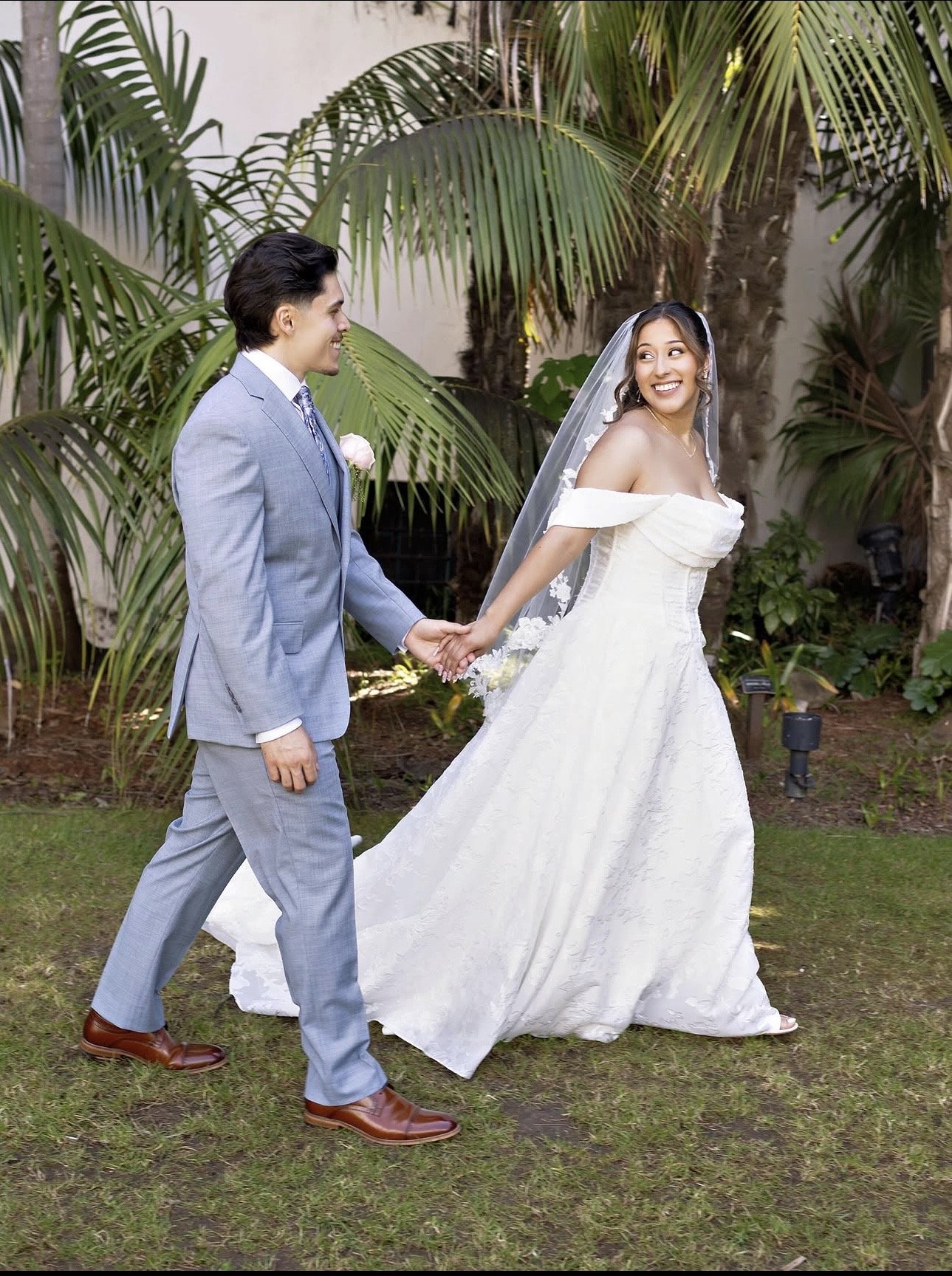A bride and groom holding hands, walking outdoors in a garden with palm trees, on their wedding day.
