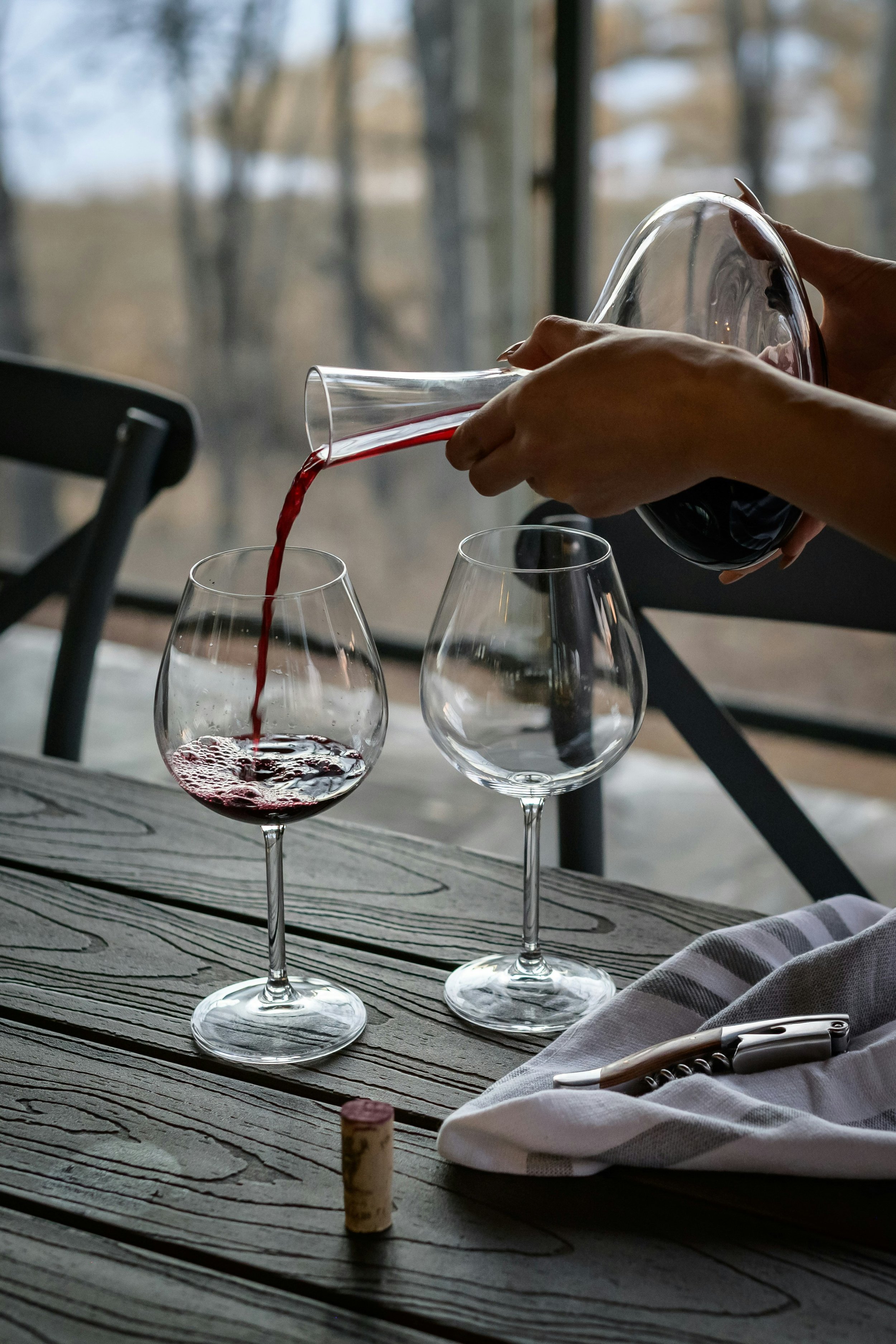 Person pouring red wine into a wine glass on a wooden table with additional empty wine glasses. There is a wine cork and a corkscrew on a napkin nearby. The background shows a window with trees outside.