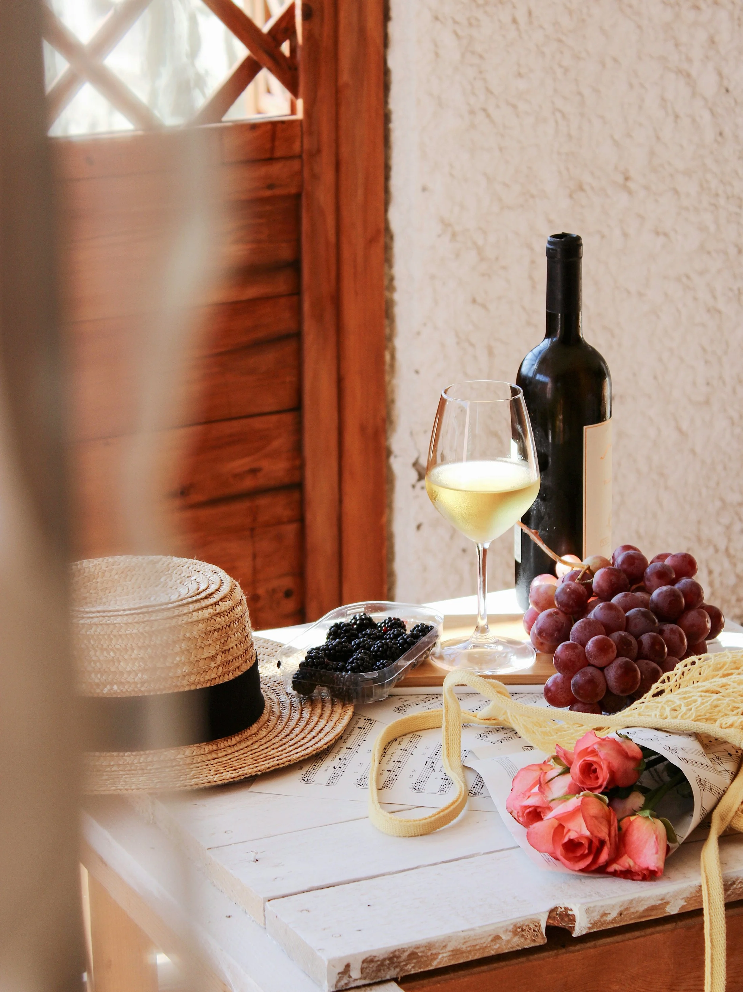 A table with a bottle of white wine, a wine glass filled with white wine, a bunch of red grapes, a small container of blackberries, a straw hat with a black band, sheet music, a bunch of pink roses, a yellow ribbon, and a beige mesh bag. The background has a textured wall and a wooden window frame.