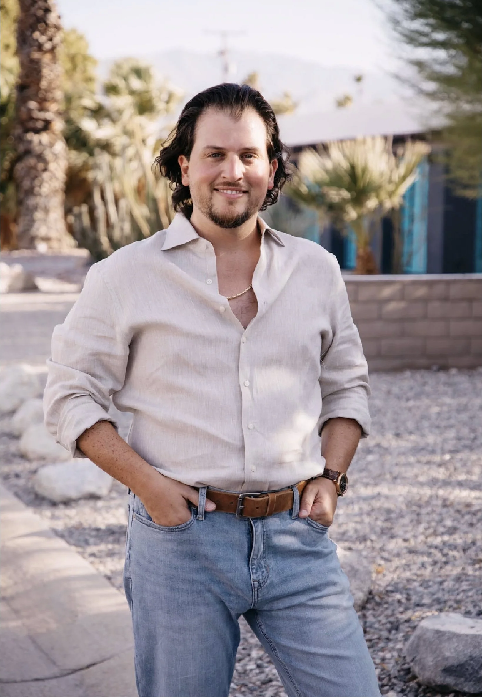 A man with dark, shoulder-length hair, a beard, and a mustache is standing outdoors, smiling. He is wearing a light-colored, long-sleeved, button-up shirt with the sleeves rolled up, blue jeans, a brown leather belt, and a watch on his left wrist. The background includes desert plants, a brick wall, and a partly cloudy sky.