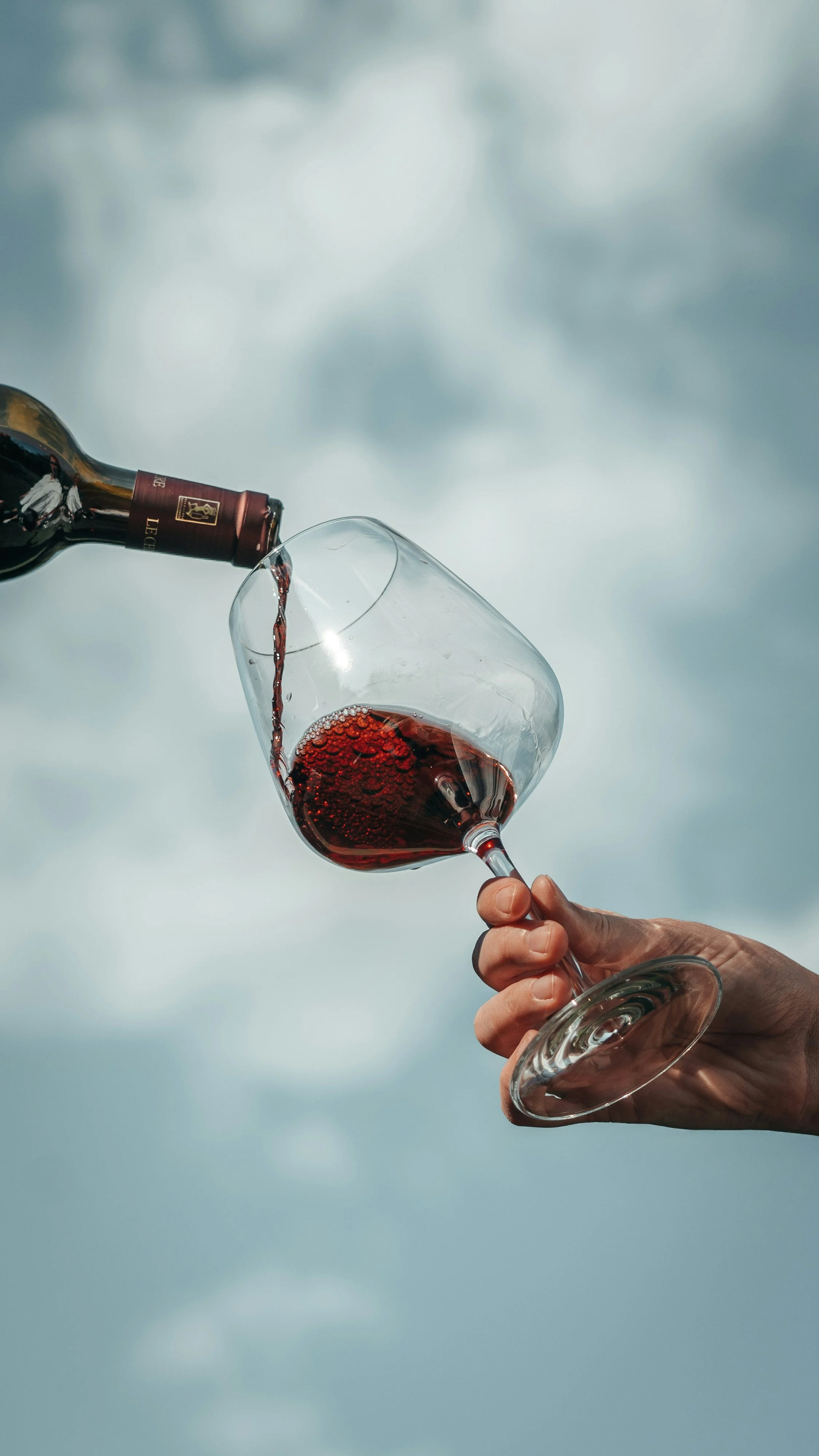Person pouring red wine into a wine glass against a cloudy sky background.