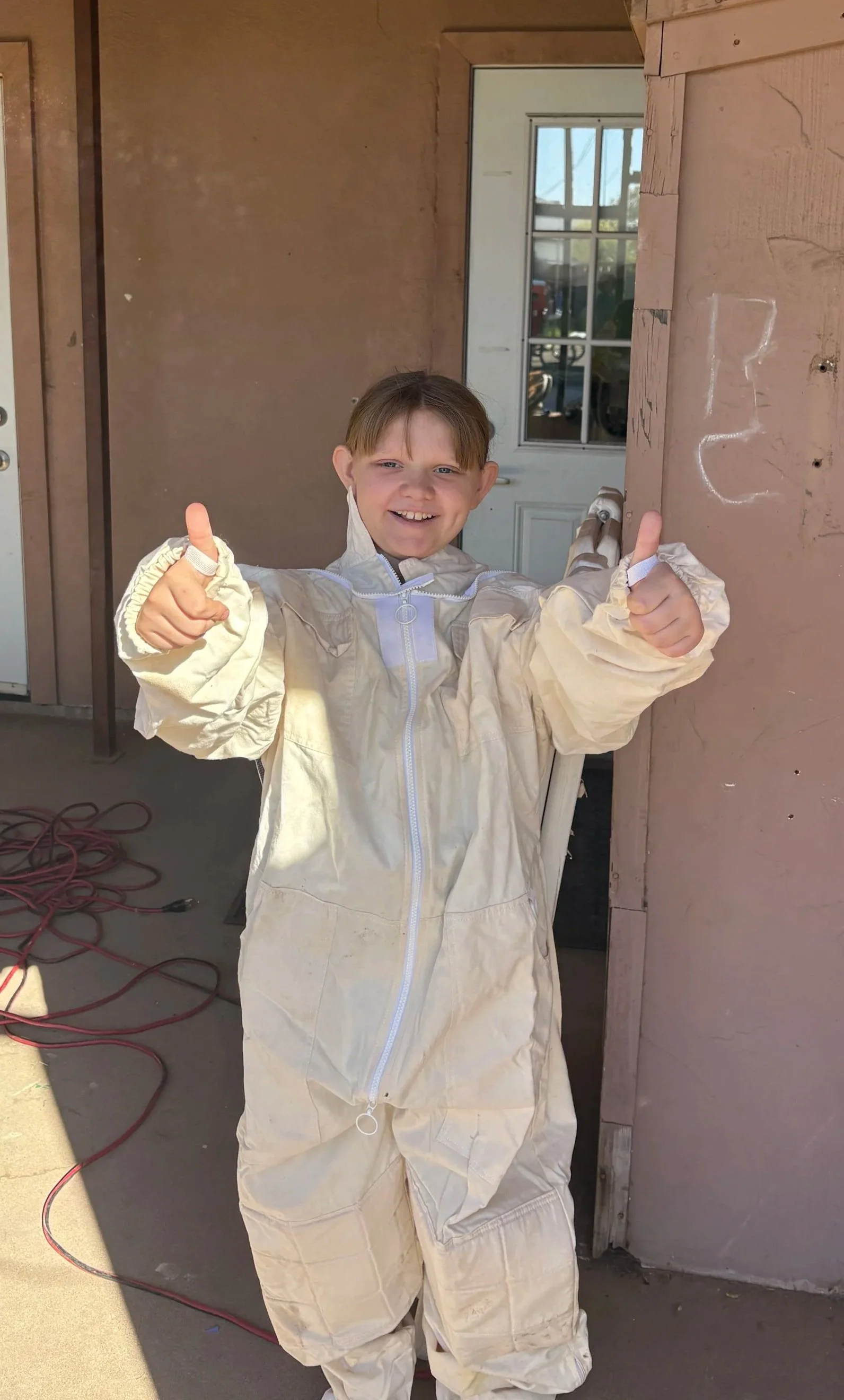 A young girl in a beige painter's jumpsuit giving two thumbs up, standing outside in front of a building with a door and window, with red extension cords on the ground.