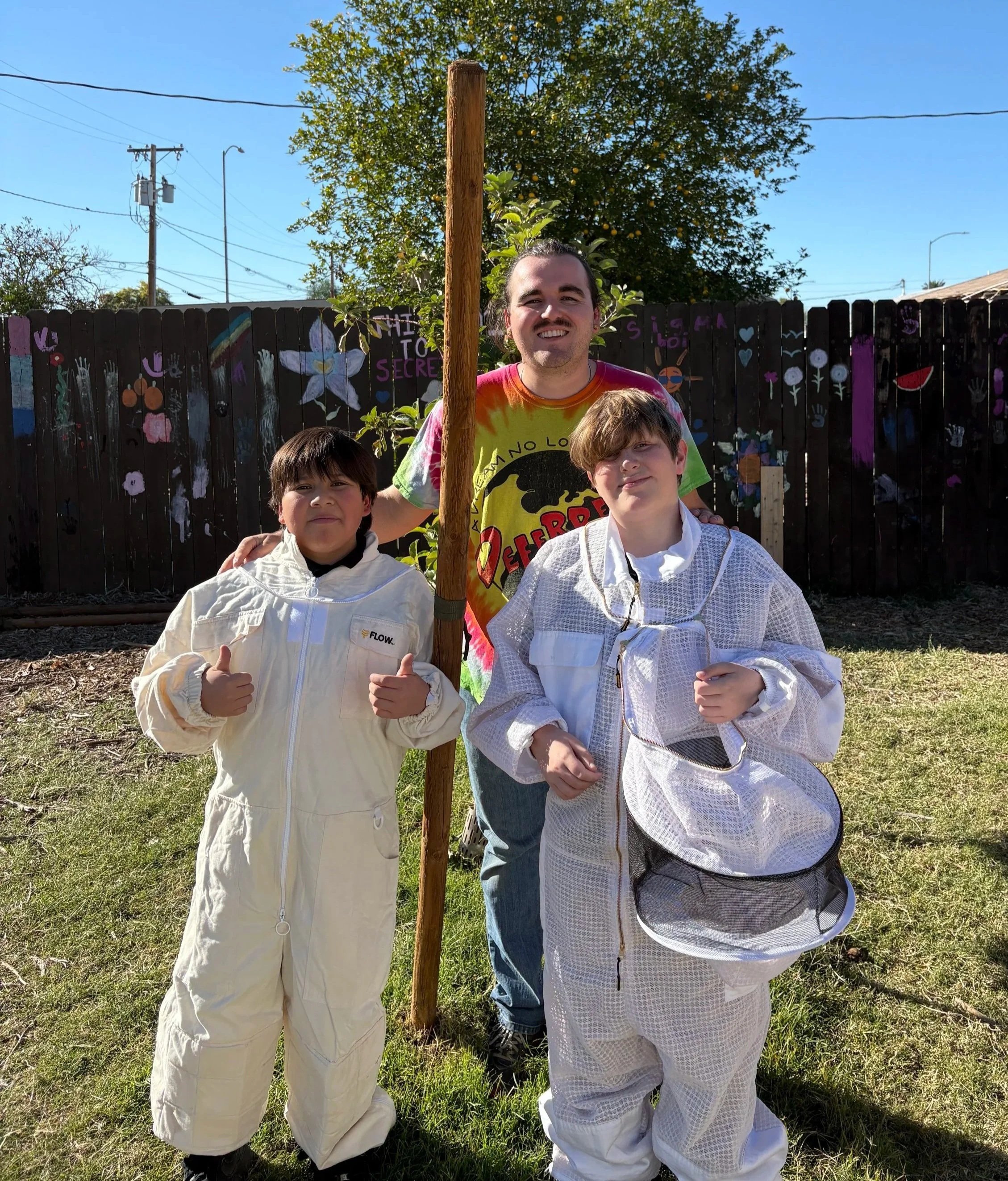 A group of three people outdoors: two children in white fencing suits and an adult male wearing a tie-dye shirt with a cartoon graphic. They are standing behind a wooden post and smiling for the photo, with a colorful decorated fence and a large lemon tree in the background.