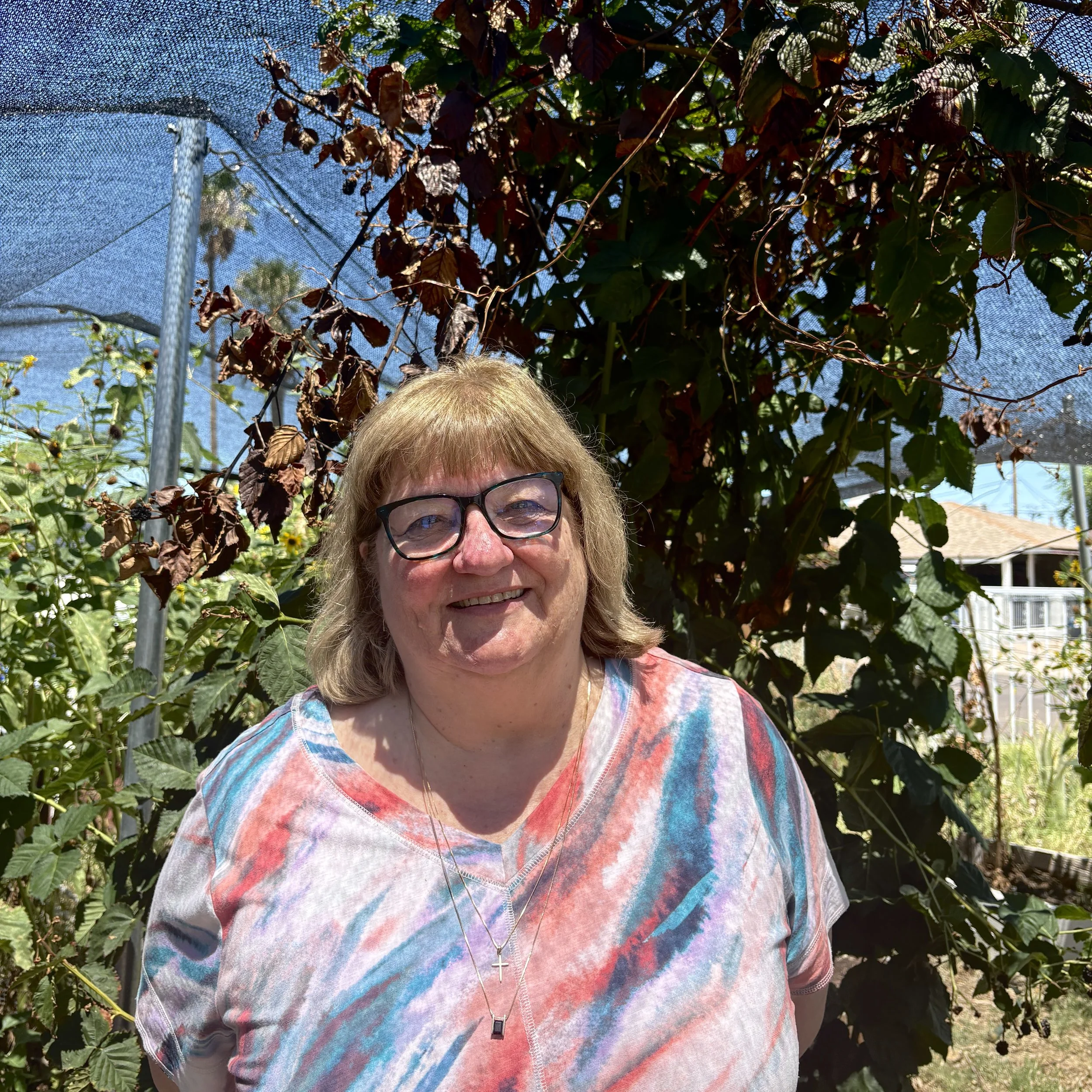 A woman with short blonde hair wearing an orange sleeveless top and beige pants sitting on a beige cushioned bench, writing in a notepad. There is a large leafy plant to her left and a wooden slatted decor on the wall. Sunlight casts shadows from a window onto the wall.