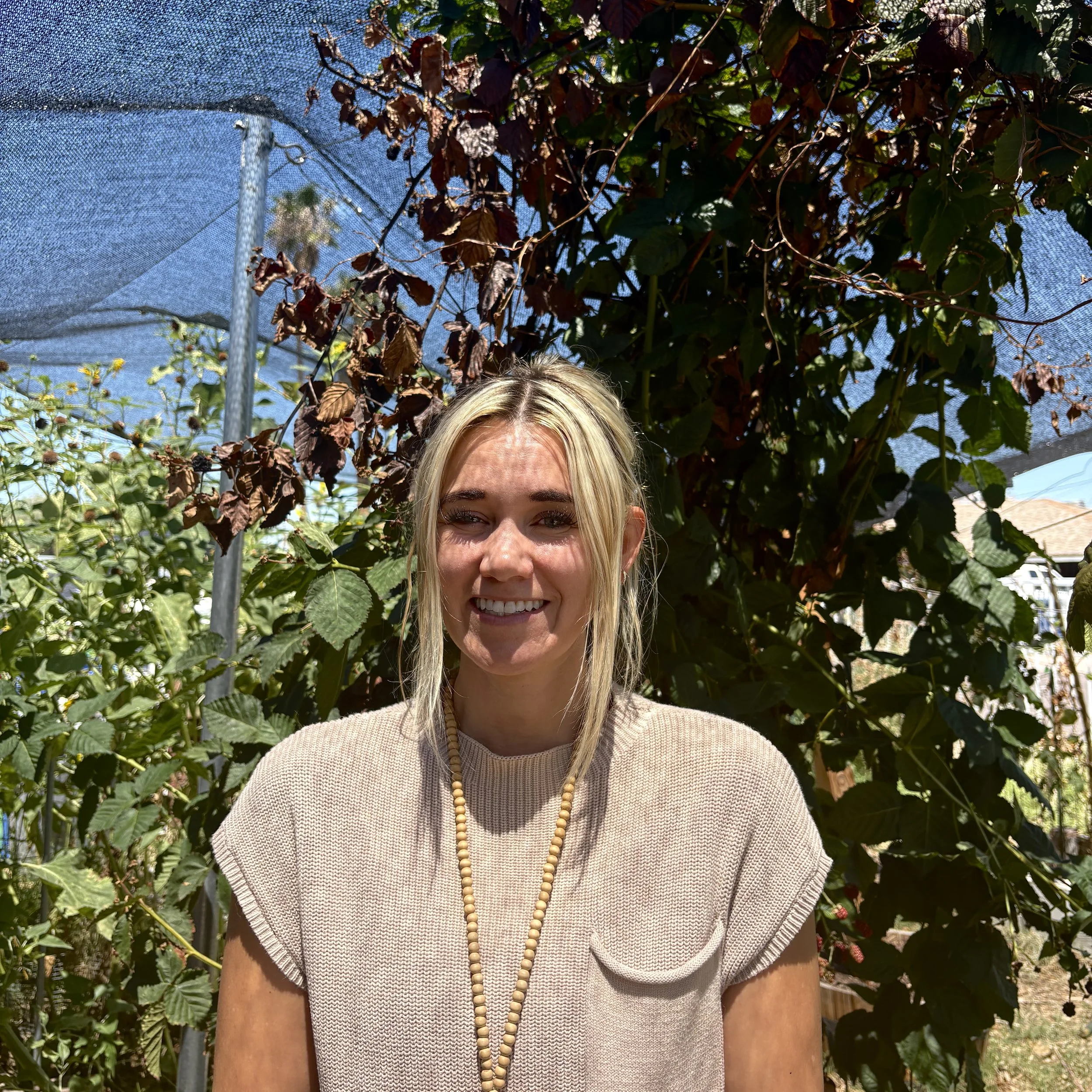 A woman with curly dark hair sitting on a beige bench, wearing a sleeveless beige top and khaki pants, in a room with a wooden slat wall and shadows cast on the wall.